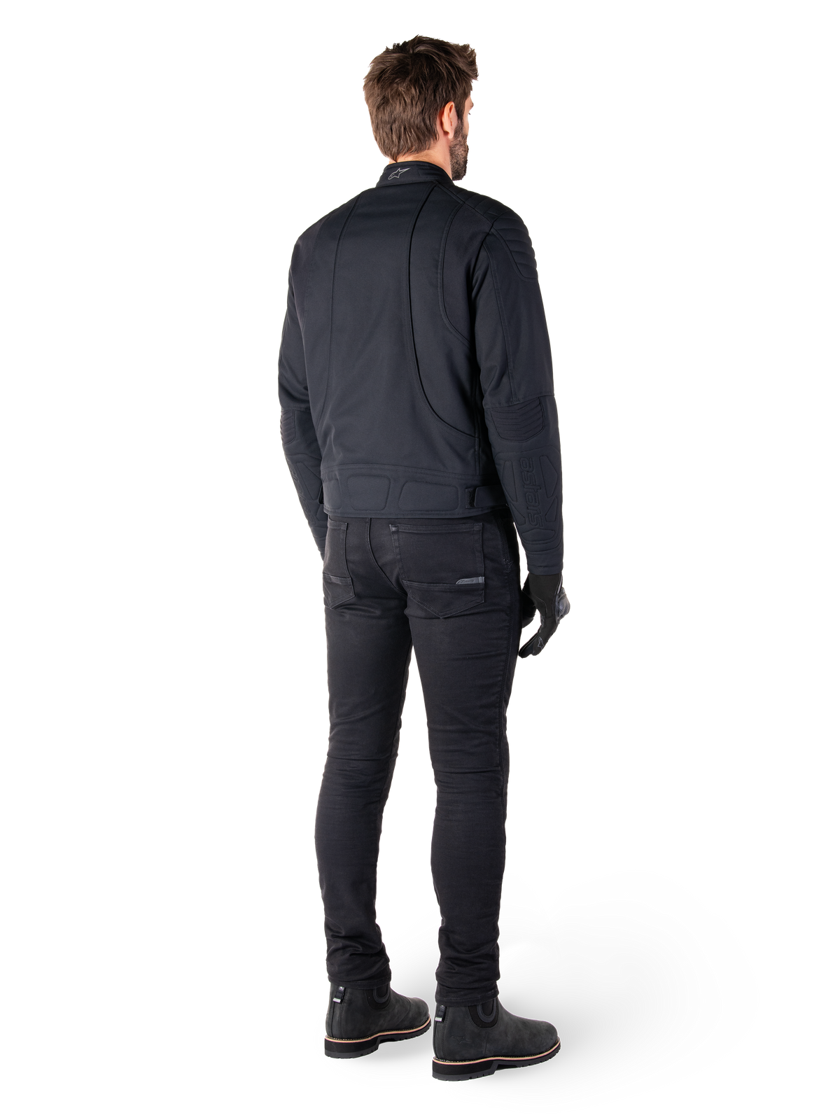 Vista posteriore di un pilota che indossa la giacca Alpinestars Clayton WR in nero, con pannelli trapuntati su gomiti e spalle, cuciture curve ergonomiche e dettagli riflettenti sul colletto, abbinata a pantaloni da moto e stivali neri per un look motociclistico urbano.