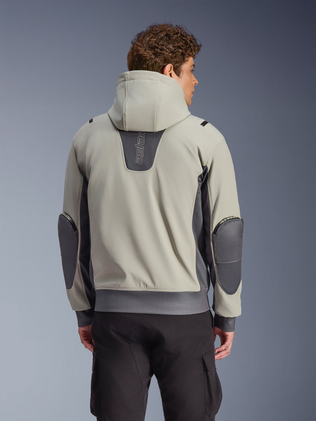 Giacca Alpinestars Moblast Waterproof in Iron Gray e Dark Sage, vista posteriore su modello, giacca da moto urbana con cappuccio dotata di pannelli elasticizzati grigio scuro e gomiti rinforzati, abbinata a pantaloni cargo neri