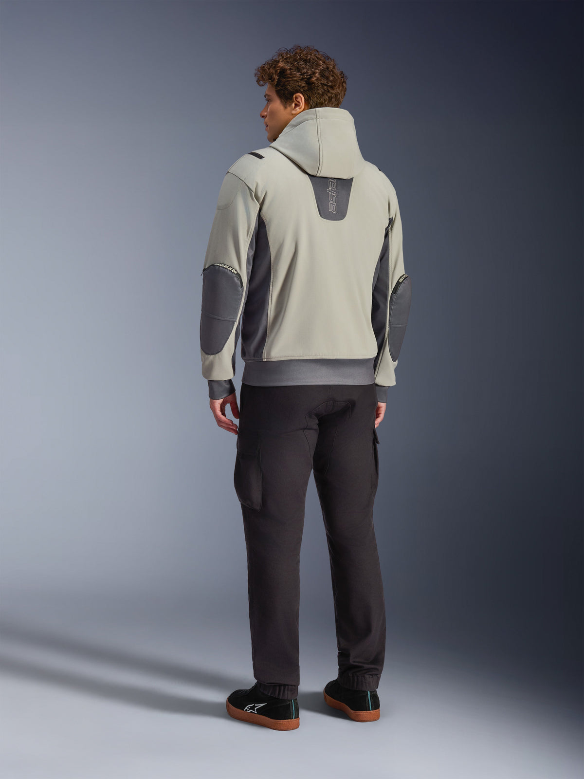 Modello visto di spalle che indossa la giacca Alpinestars Moblast Waterproof in Iron Gray Dark Sage, giacca da moto urbana con cappuccio con pannelli laterali grigio scuro e toppe rinforzate sui gomiti, abbinata a pantaloni da guida scuri e sneaker nere, estetica moderna e minimalista per il commuting urbano.