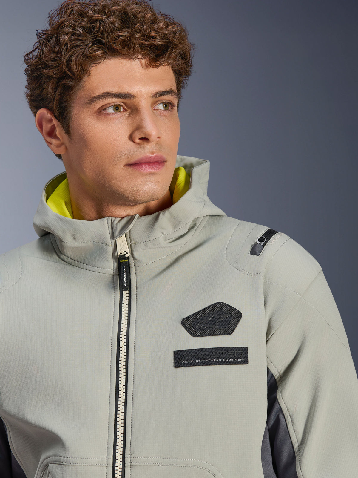 Giacca impermeabile Alpinestars Moblast, Iron Gray Dark Sage, primo piano di un modello maschile che indossa la giacca, con logo Alpinestars nero e patch MO.ST.EQ. sul petto, fodera del cappuccio giallo brillante e design tecnico moderno.