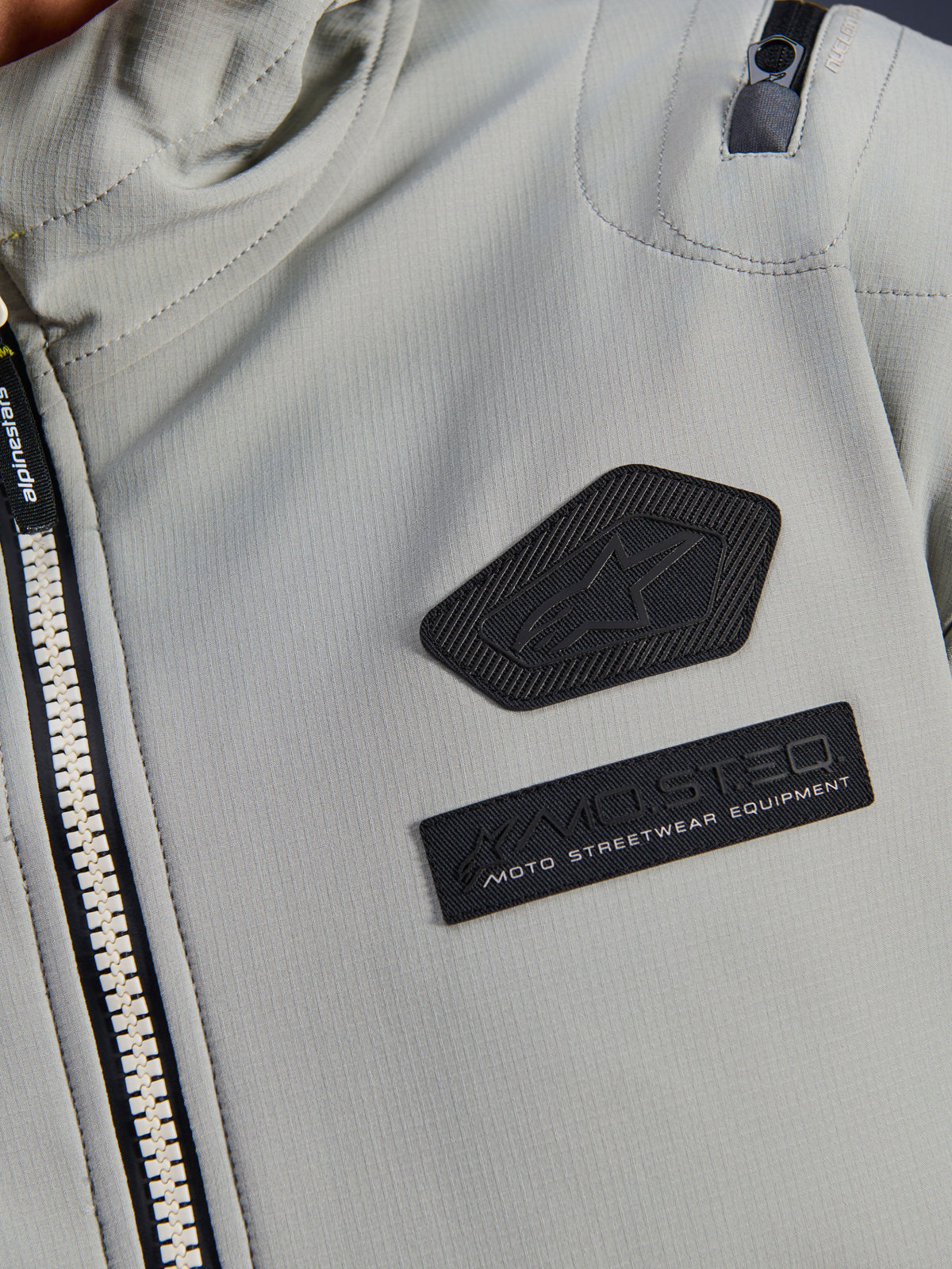 Giacca impermeabile Alpinestars Moblast, Iron Gray, dettaglio ravvicinato delle patch sul petto con logo Alpinestars e marchio MO.ST.EQ. Moto Streetwear Equipment, tessuto tecnico ripstop con robusta cerniera frontale e dettaglio della tasca sulla spalla.