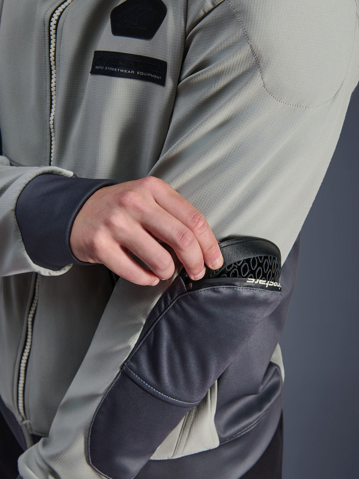 Alpinestars Moblast Waterproof Jacket, Iron Gray e Dark Sage, primo piano che mostra la rimozione della protezione Nucleon Flex Plus dalla tasca esterna con cerniera sul gomito, abbigliamento tecnico da moto per uso urbano con marchio MO.ST.EQ.