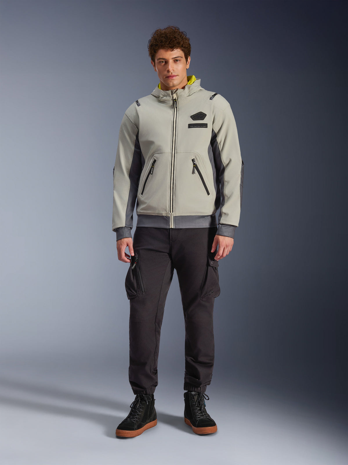 Modello che indossa la giacca Alpinestars Moblast Waterproof in Iron Gray e Dark Sage, giacca da moto con cappuccio dal design tecnico moderno, abbinata a pantaloni cargo da moto scuri e stivali neri, abbigliamento motorsport urbano contemporaneo