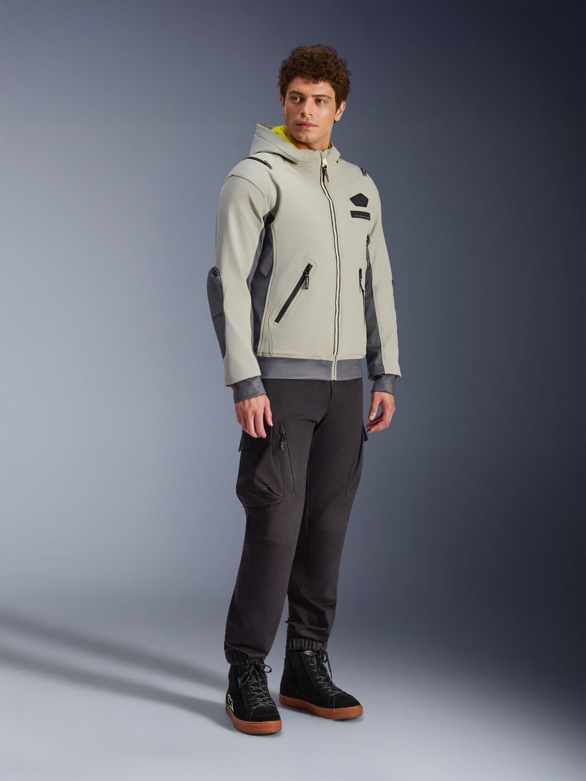 Alpinestars Moblast Waterproof Jacket, Iron Gray e Dark Sage, giacca da moto urbana con design a felpa con cappuccio, indossata da un modello con pantaloni da moto scuri e stivali neri, estetica cyber-tech minimalista con interno del cappuccio giallo e cerniere impermeabili