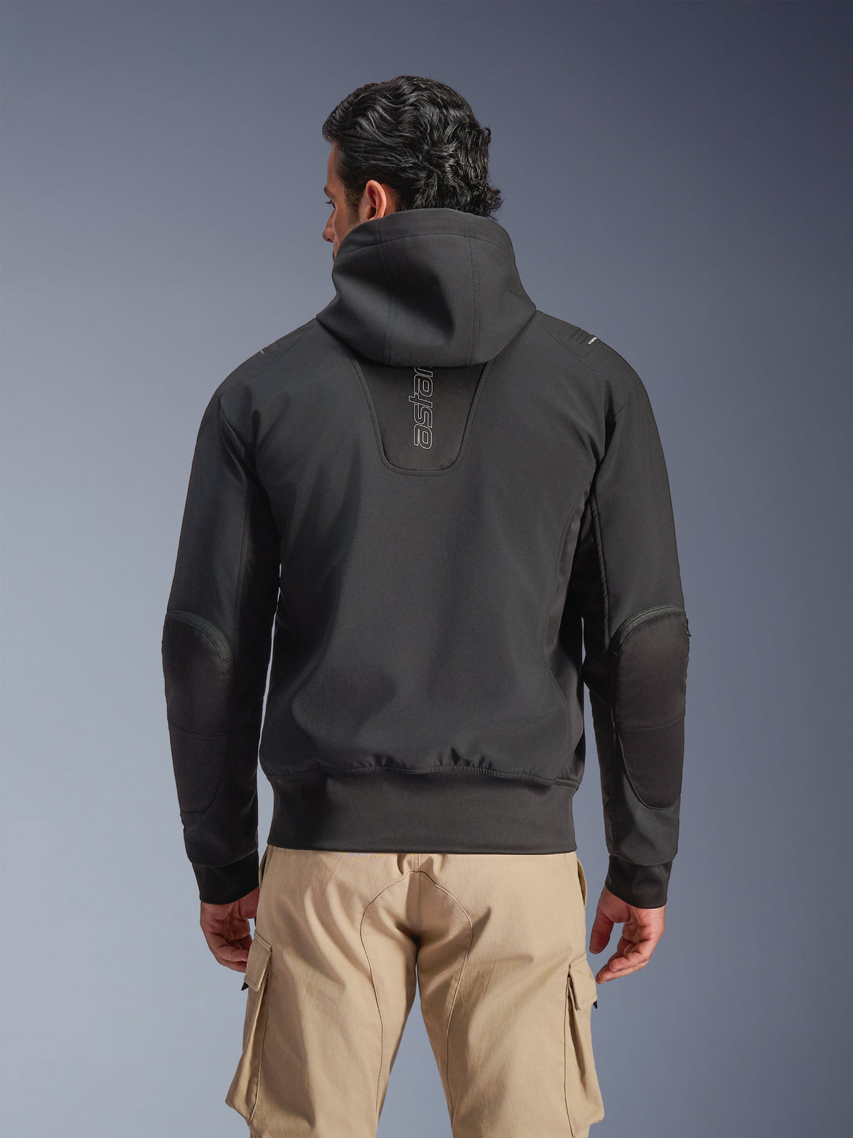 Alpinestars Giacca Moblast Waterproof, Nero, vista posteriore di giacca da moto urbana con cappuccio, con logo 'astars' verticale sulla schiena, vita e polsini elasticizzati, gomiti rinforzati e design moderno e tecnologico, indossata con pantaloni cargo beige