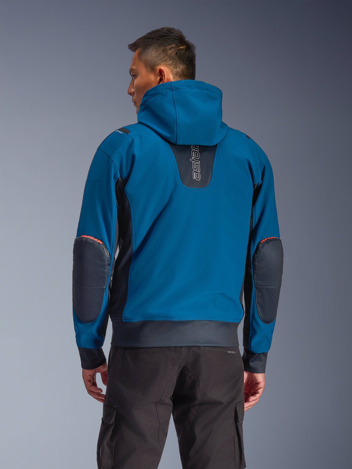 Giacca impermeabile Alpinestars Moblast, colori Dark Petrol e Dark Navy, felpa con cappuccio da moto urbana vista dal retro, con cappuccio fisso, logo 'astars' verticale sulla parte superiore della schiena, toppe rinforzate scure sui gomiti, vita e polsini elasticizzati, abbigliamento tecnico moderno da moto