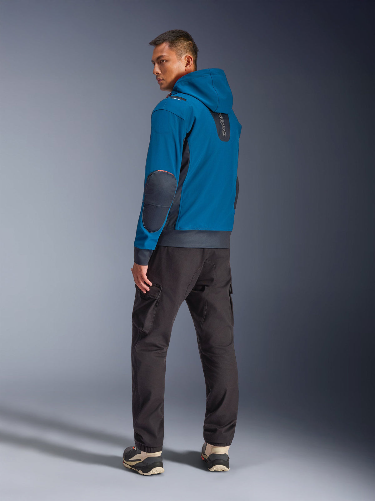 Alpinestars Moblast Waterproof Jacket in Dark Petrol e Dark Navy, vista posteriore su modello che mostra il design con cappuccio, toppe sui gomiti e inserti laterali in dark navy, abbinata a pantaloni cargo scuri e sneakers tecniche, abbigliamento da moto urbano moderno.
