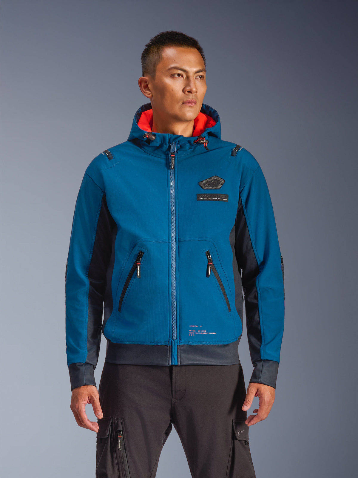 Alpinestars Moblast Waterproof Jacket indossata da un modello, blu e blu navy, giacca da moto urban riding, design con cappuccio e interno rosso, cerniere nere e dettaglio patch sul petto, abbigliamento da moto ispirato allo streetwear
