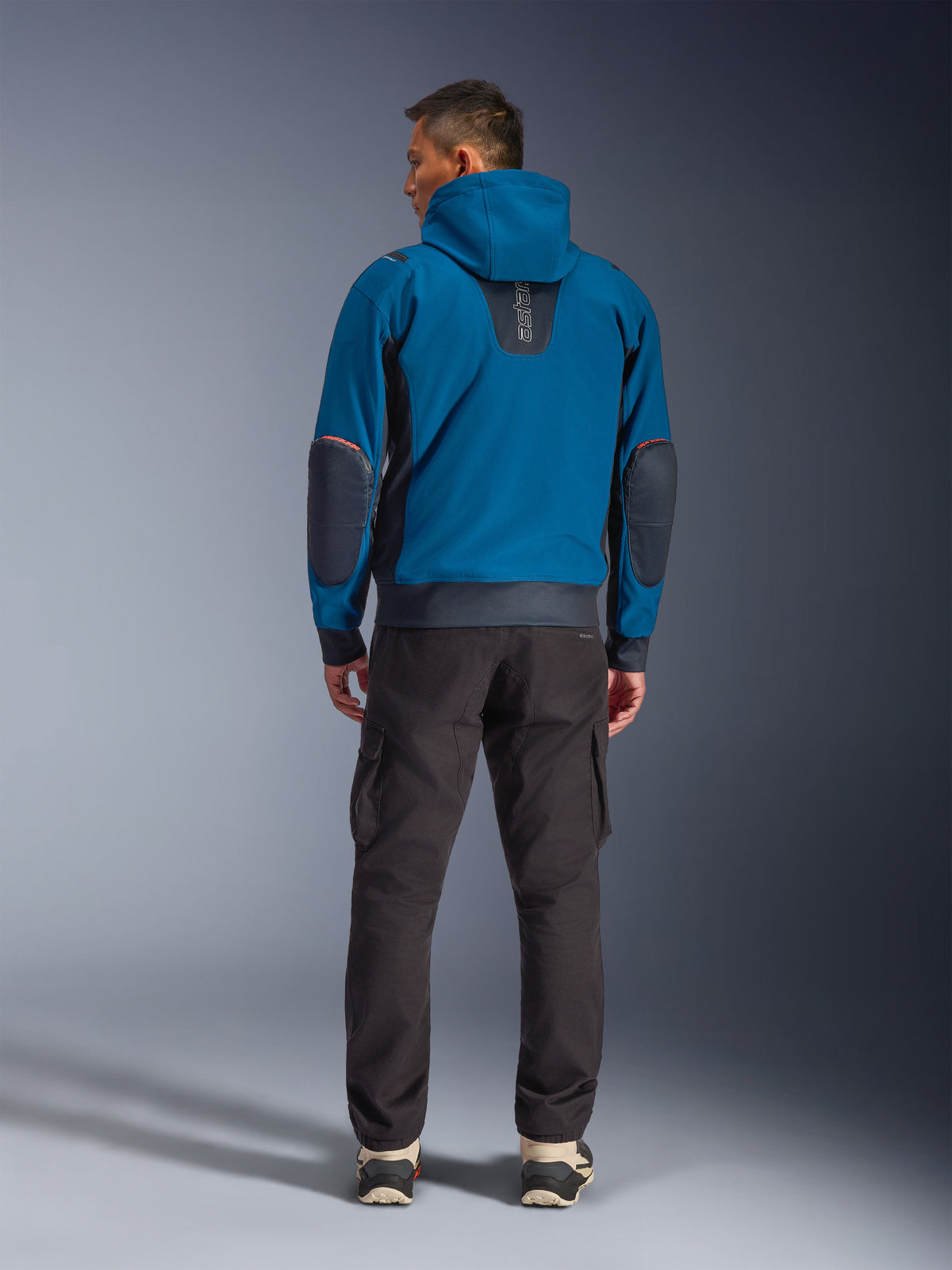 Giacca impermeabile Alpinestars Moblast, giacca da moto urbana, Dark Petrol e Dark Navy Blue, vista posteriore con cappuccio e toppe rinforzate sui gomiti, indossata con pantaloni cargo scuri, abbigliamento tecnico elegante da commuter per tutte le stagioni