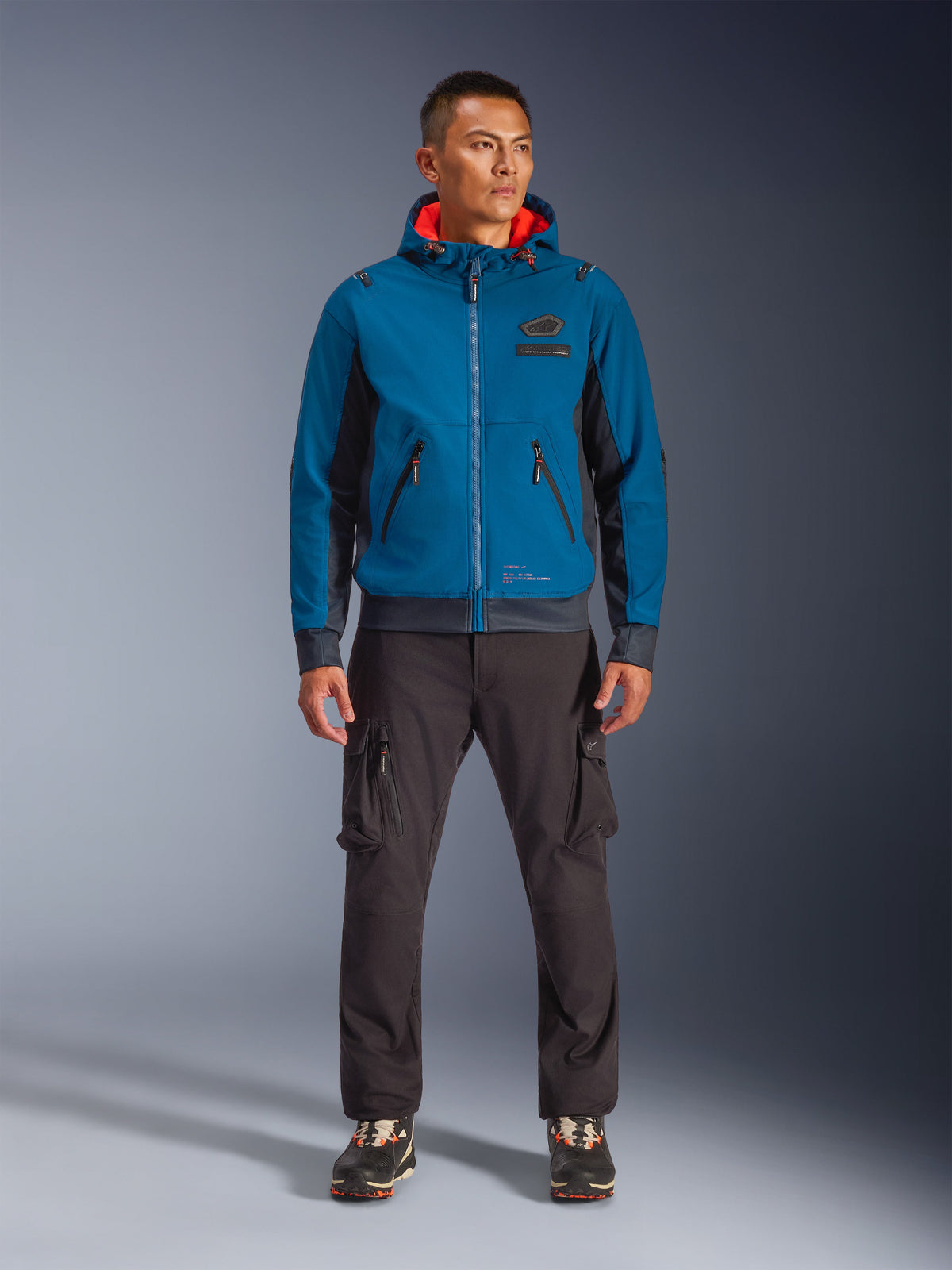 Alpinestars Moblast Waterproof Jacket, giacca da moto con cappuccio in blu petrolio scuro e blu navy con fodera interna arancione, indossata da un modello con pantaloni cargo scuri e sneakers nere, abbigliamento moderno da guida urbana