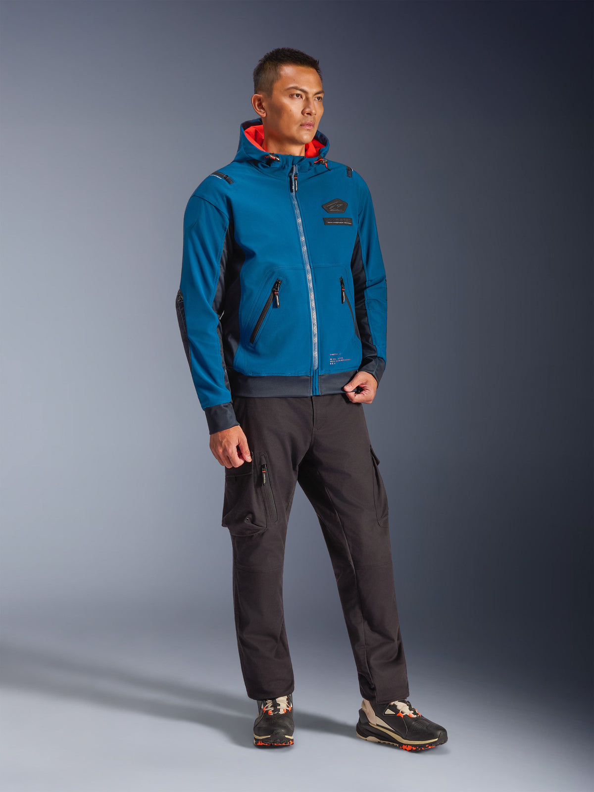 Modello che indossa la giacca Alpinestars Moblast Waterproof in Dark Petrol e Dark Navy, design con cappuccio e interno arancione acceso, abbinata a pantaloni cargo neri e sneakers, abbigliamento tecnico da moto urbano moderno.