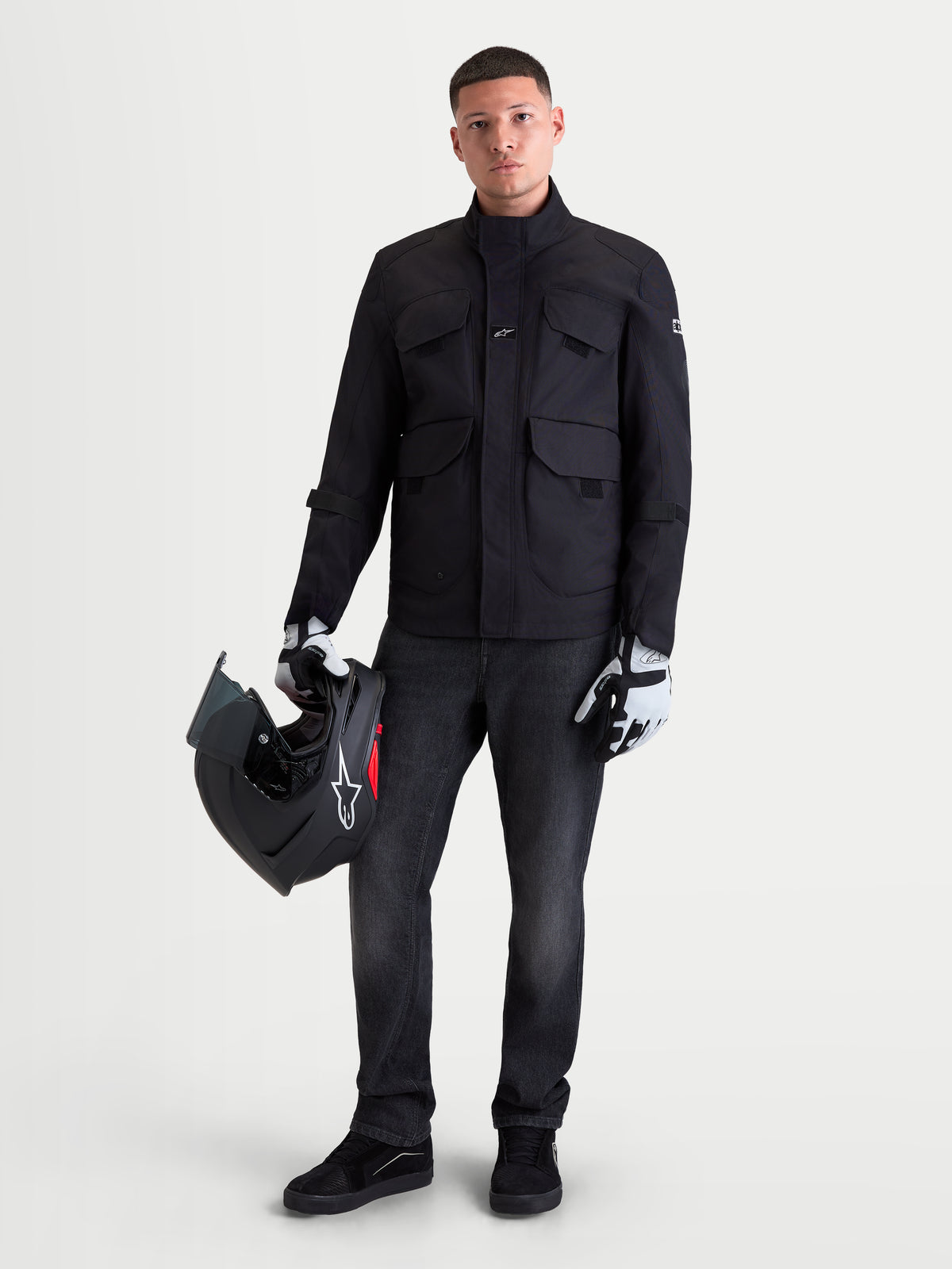 Alpinestars Altan Drystar Giacca, giacca da moto urbana contemporanea da uomo in nero, vista frontale su un modello che tiene un casco e indossa guanti da moto, giacca utility in tessuto impermeabile con tasche cargo