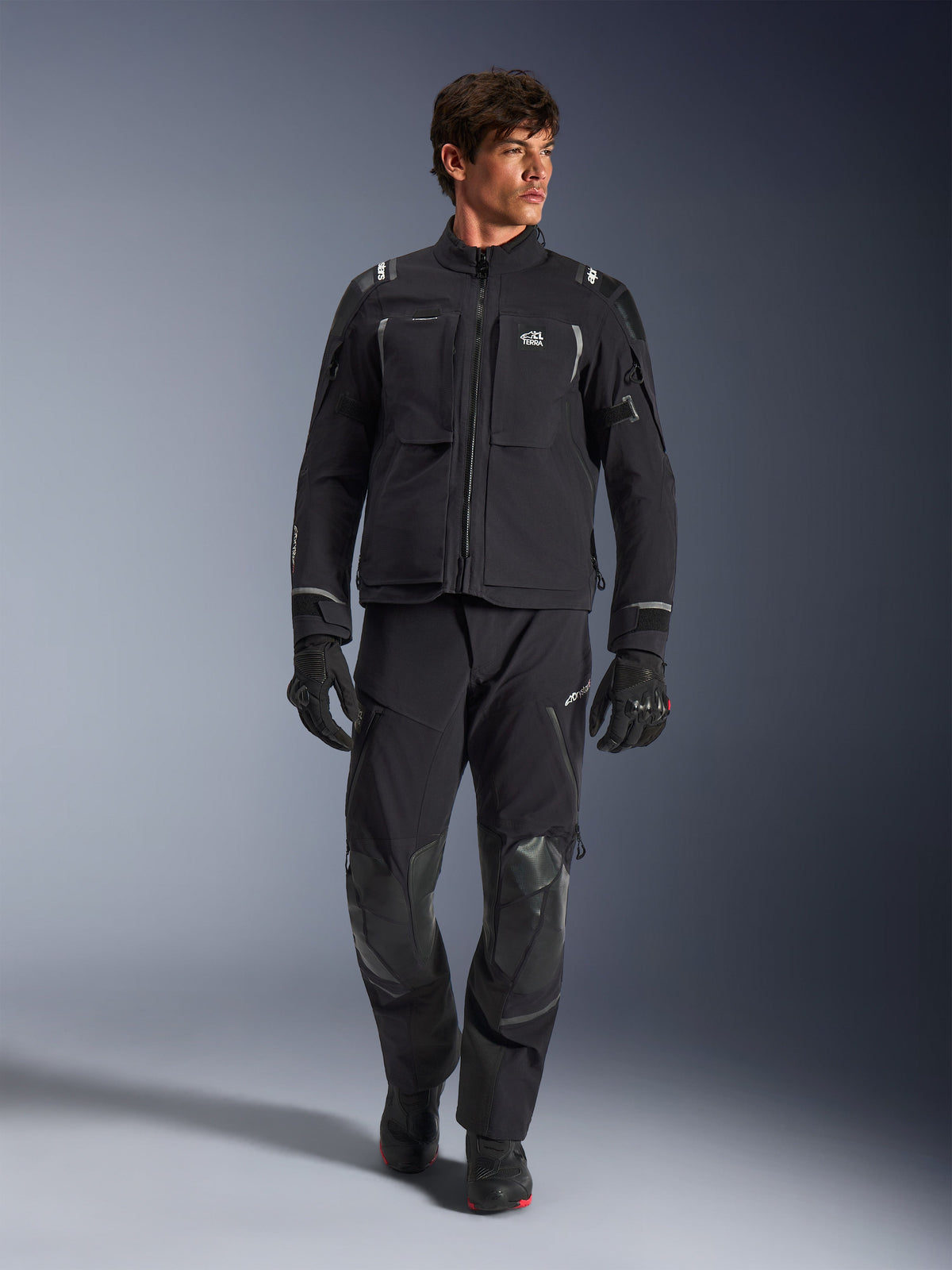 Modello che indossa la giacca Alpinestars Andes Pro Drystar® XF, giacca da moto adventure in nero e grigio scuro, abbinata a pantaloni tecnici e guanti coordinati, con tasche multiple e marchio 'Terra' sul petto.