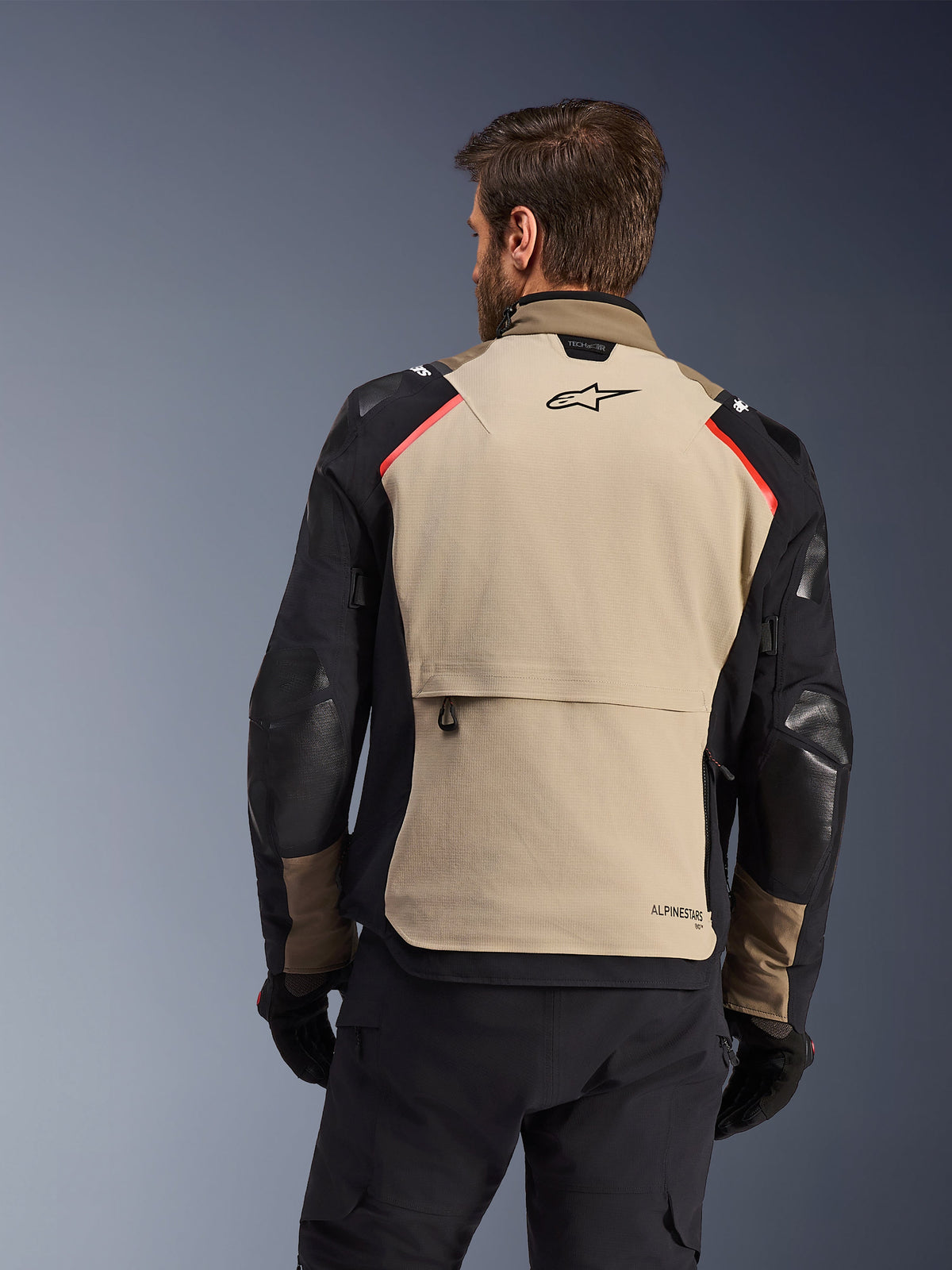 Giacca Alpinestars Andes Pro Drystar® XF, vista posteriore, color noce e alluminio, con un ampio pannello posteriore color sabbia, logo Alpinestars nero e accenti rossi sulle spalle, grande tasca cargo posteriore, indossata con pantaloni e guanti neri, abbigliamento da moto adventure touring