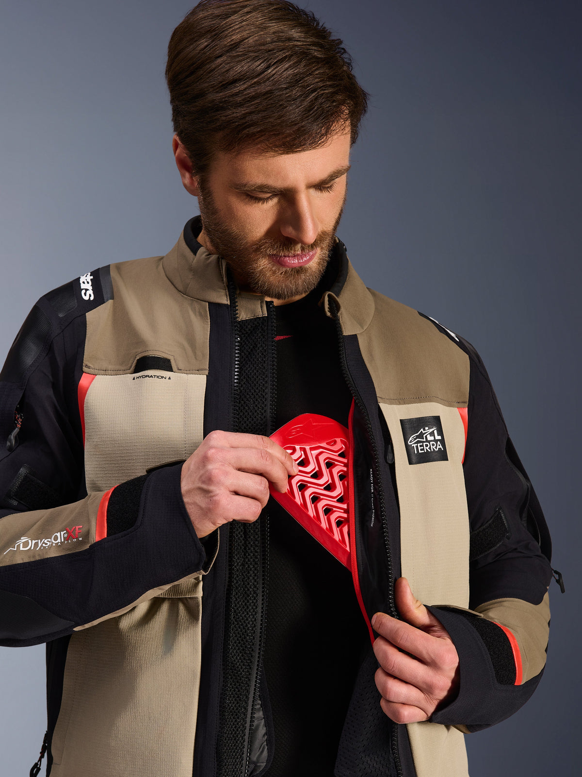 Giacca Alpinestars Andes Pro Drystar® XF in colorazione marrone noce e alluminio, modello che mostra l'inserimento della protezione per il torace rossa nella tasca interna, giacca da moto adventure con marchio ALL TERRA e loghi Drystar XF