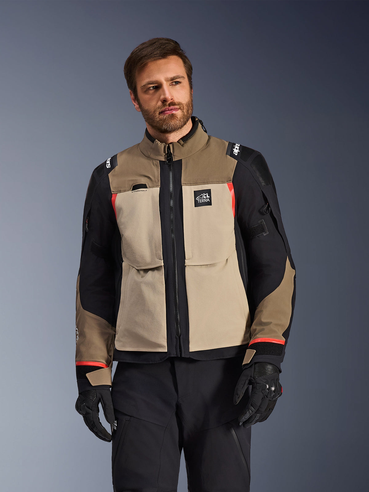 Giacca Alpinestars Andes Pro Drystar® XF, Walnut Aluminum, giacca da moto adventure con pannelli color sabbia e neri e accenti rossi, indossata con pantaloni da guida e guanti neri, abbigliamento tecnico da touring impermeabile