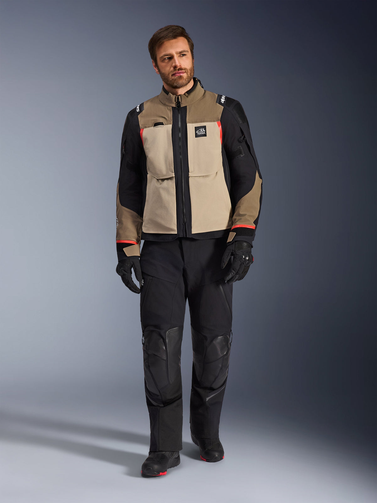 Modello con giacca Alpinestars Andes Pro Drystar® XF in Walnut Aluminum, giacca da touring adventure marrone e nera con accenti rossi, abbinata a pantaloni da moto, guanti e stivali neri, scatto in studio a figura intera per abbigliamento protettivo.