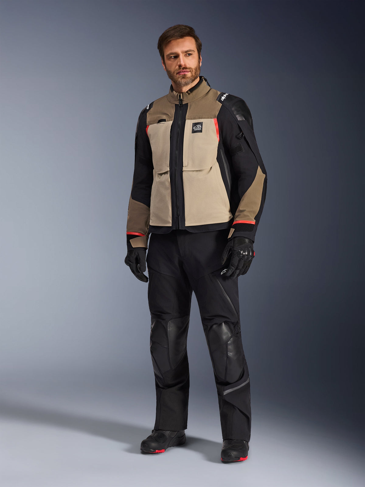 Un modello maschile che indossa la giacca Alpinestars Andes Pro Drystar® XF in Walnut Aluminum, abbinata a pantaloni da moto, guanti e stivali neri, caratterizzata da un design multi-pannello beige e nero con accenti rossi per adventure touring.