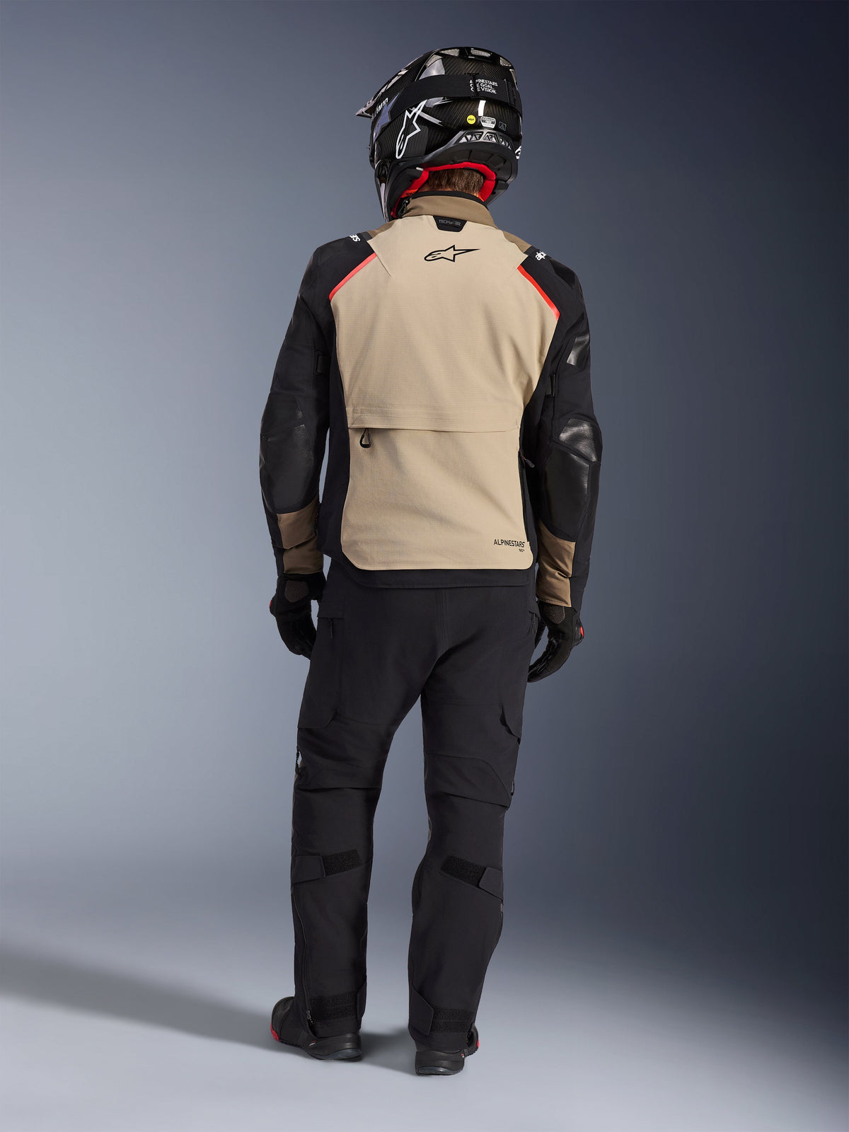 Pilota visto da dietro che indossa la giacca Alpinestars Andes Pro Drystar® XF in Walnut Aluminum, pannello posteriore color sabbia con maniche nere e dettagli rossi, abbinata a pantaloni tecnici adventure neri e casco da moto, equipaggiamento touring premium per tutte le stagioni