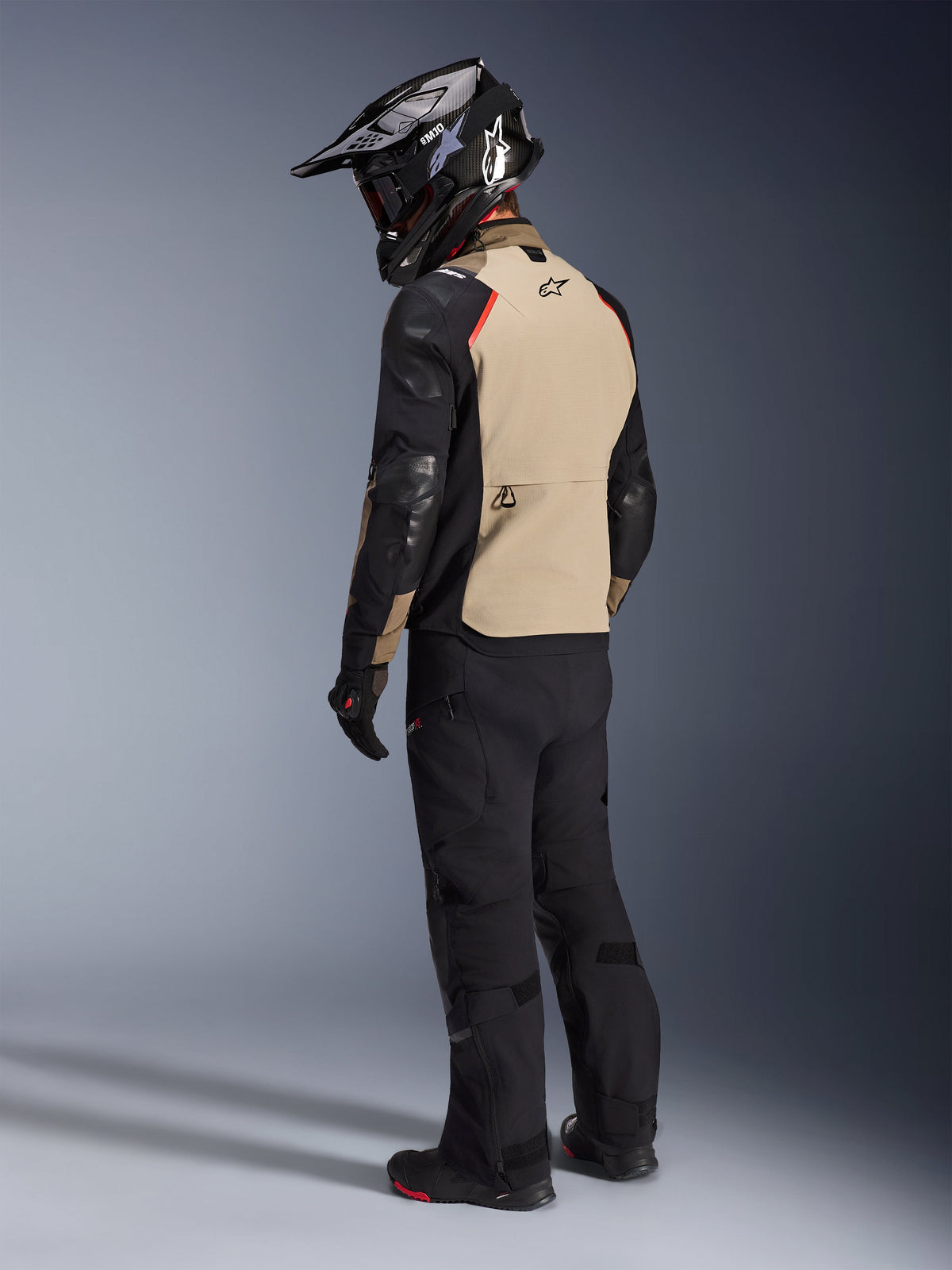 Motociclista visto da dietro con la giacca Alpinestars Andes Pro Drystar® XF in Walnut Aluminum, con pannello posteriore color sabbia, maniche nere e dettagli rossi, casco adventure nero, pantaloni da moto neri e stivali neri, abbigliamento professionale da touring adventure.