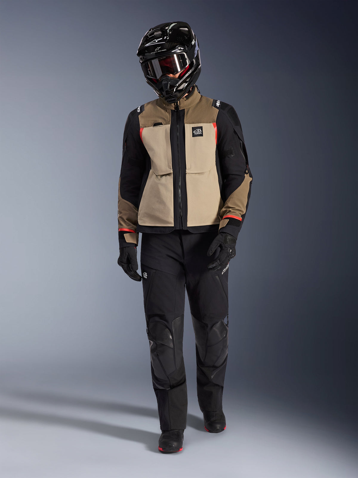Giacca Alpinestars Andes Pro Drystar® XF in Walnut Aluminum, indossata con pantaloni adventure neri, guanti neri e casco integrale nero con maschera, con dettagli rossi e marchio All Terra, set-up professionale per adventure touring