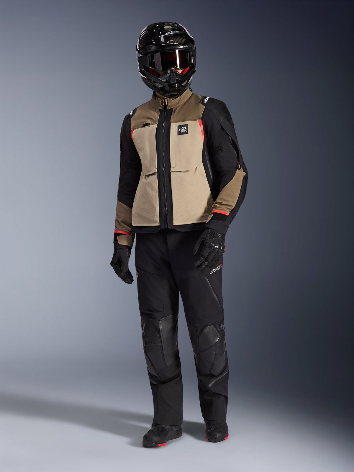 Alpinestars Andes Pro Drystar® XF Jacket in Walnut Aluminum, giacca da moto adventure indossata con pantaloni neri abbinati, casco adventure nero con maschera e stivali neri, caratterizzata da pannelli color sabbia e neri con dettagli rossi.