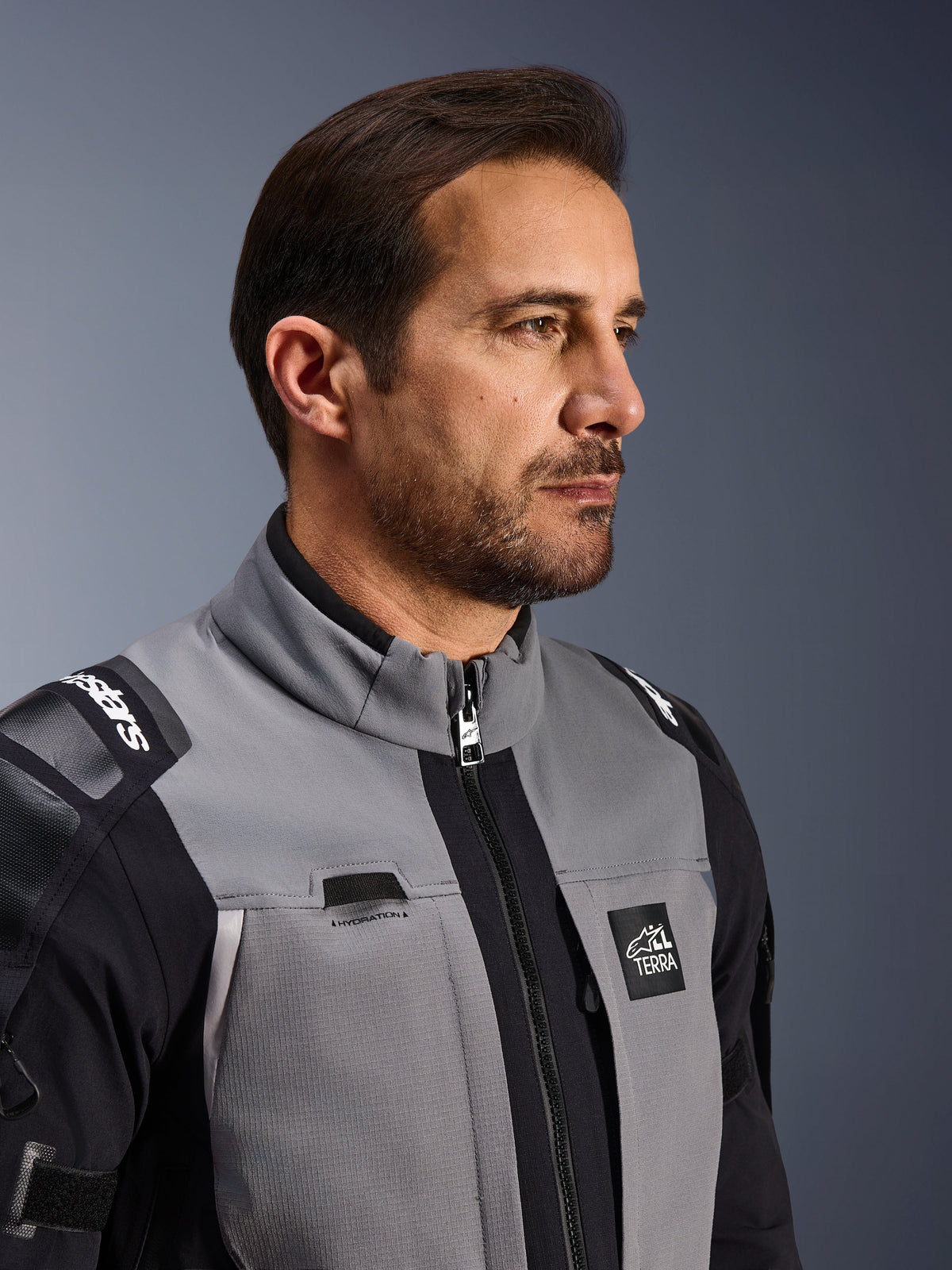 Giacca Alpinestars Andes Pro Drystar® XF, giacca da moto adventure, grigio scuro e nero, primo piano della parte anteriore del busto e del colletto, con logo Terra e dettaglio del passaggio per l'idratazione, indossata da un uomo in uno studio.