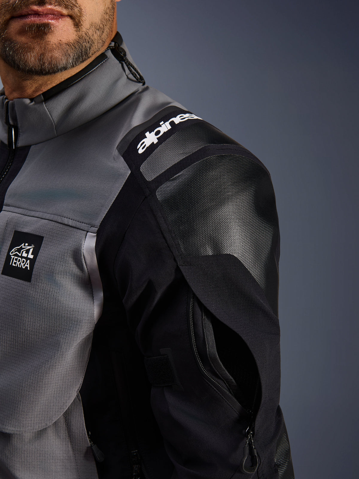 Giacca Alpinestars Andes Pro Drystar® XF, giacca da moto adventure, grigio scuro e nero, vista ravvicinata della spalla rinforzata con marchio Alpinestars, patch con logo 'TERRA' e ventilazione della manica con zip su guscio in tessuto tecnico.