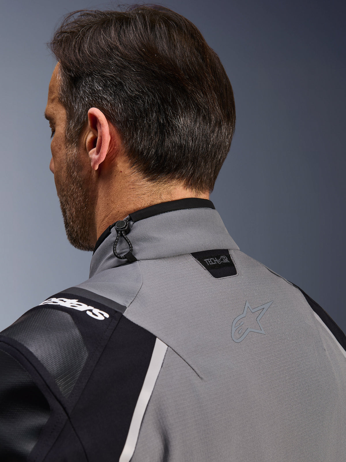 Giacca Alpinestars Andes Pro Drystar® XF, grigio scuro e nero, primo piano del colletto posteriore e della parte superiore della spalla, mostra il logo Tech-Air® Ready, la regolazione con cordino elastico e il logo a stella Alpinestars in tinta su tessuto tecnico