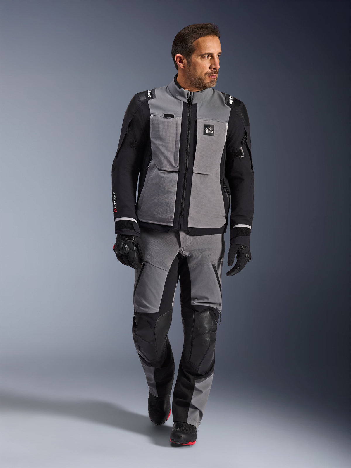 Modello che indossa la giacca Alpinestars Andes Pro Drystar® XF in grigio scuro e nero, completo da moto adventure con pantaloni e guanti coordinati, guscio in tessuto tecnico con marchio 'All Terra', equipaggiamento completo per il touring adventure a lunga distanza
