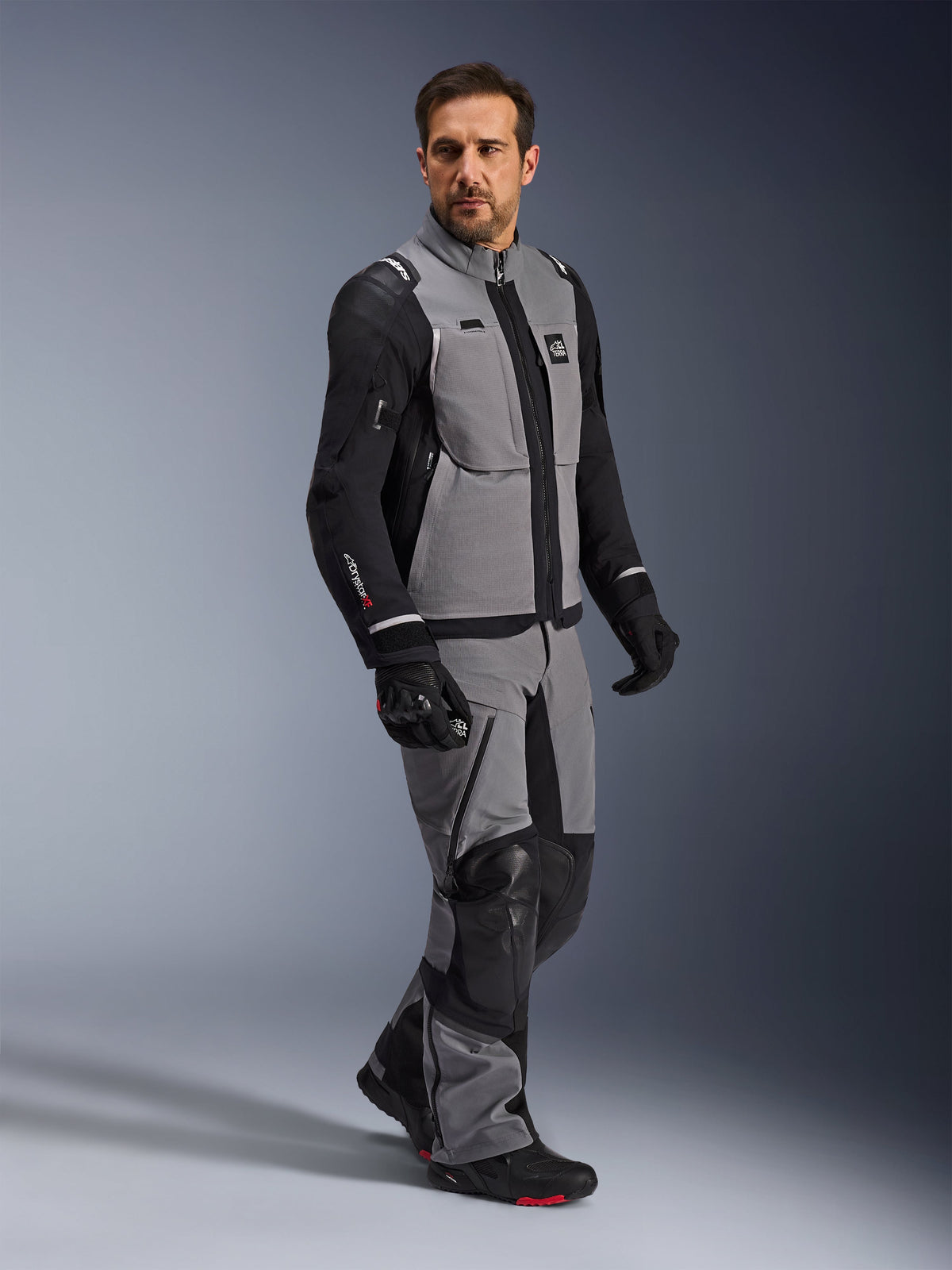 Giacca Alpinestars Andes Pro Drystar® XF, giacca da moto adventure, grigio scuro e nero, indossata da un modello con pantaloni in tessuto coordinati e guanti neri, caratterizzata da un design multitasche, guscio laminato tecnico a 3 strati e vestibilità articolata per viaggi adventure a lunga distanza.