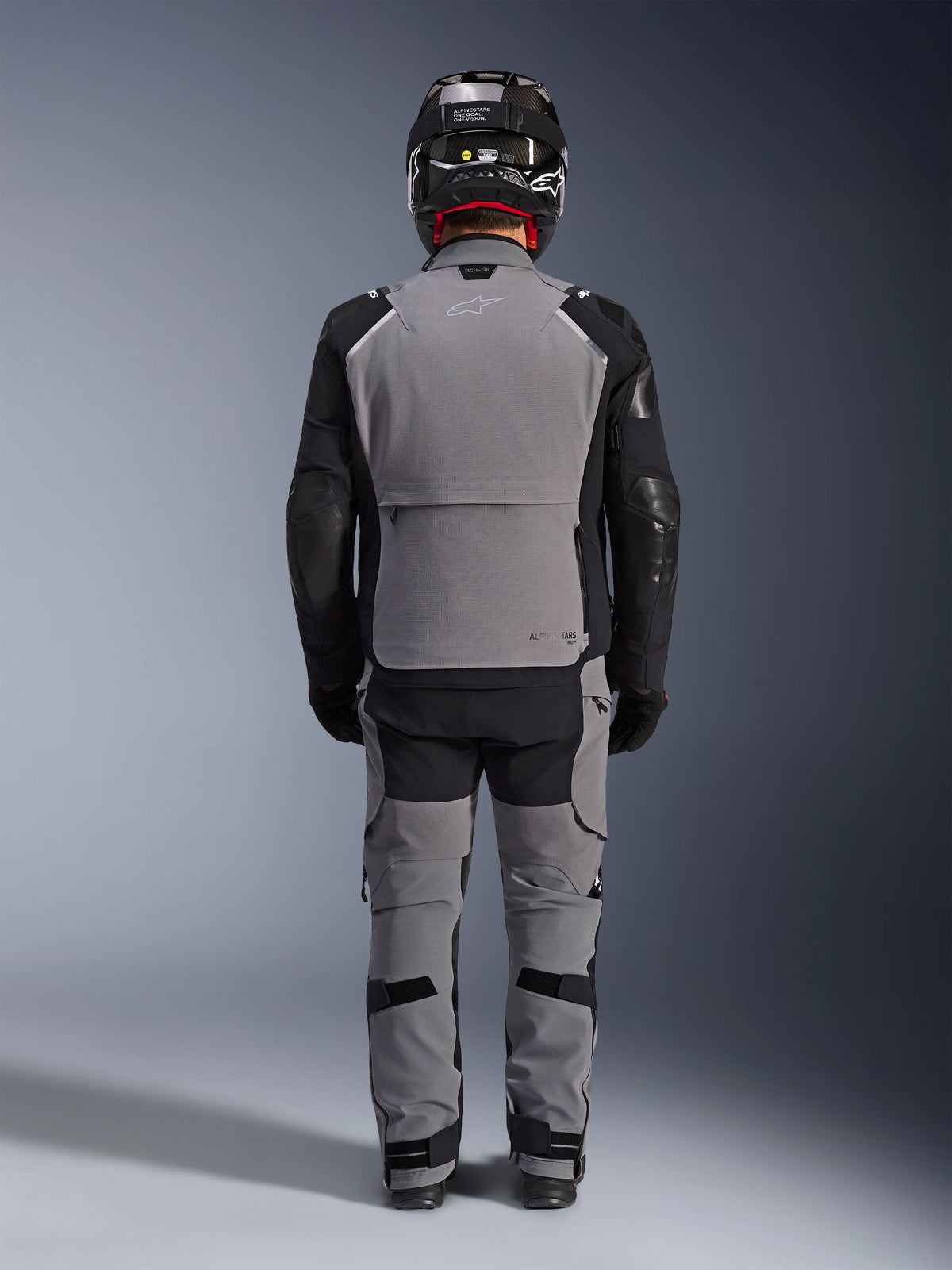 Giacca Alpinestars Andes Pro Drystar® XF, vista posteriore, abbigliamento adventure touring grigio scuro e nero, indossata con pantaloni coordinati grigi e neri, casco in fibra di carbonio nero e guanti neri, con pannelli tecnici e loghi riflettenti