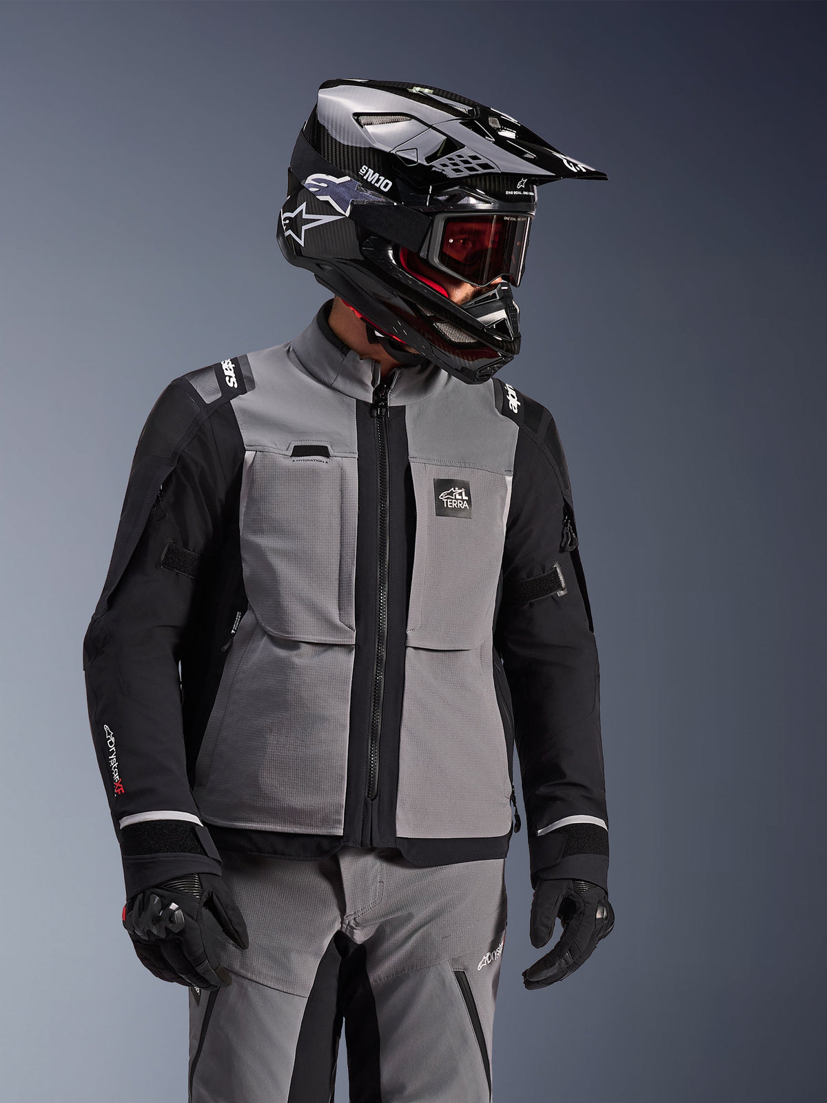 Giacca Alpinestars Andes Pro Drystar® XF, grigio scuro e nero, giacca da moto adventure indossata con casco SM10 nero e abbigliamento tecnico coordinato, design in tessuto a 3 strati con cerniera centrale e tasche sul petto, design articolato per il touring in ogni condizione climatica.