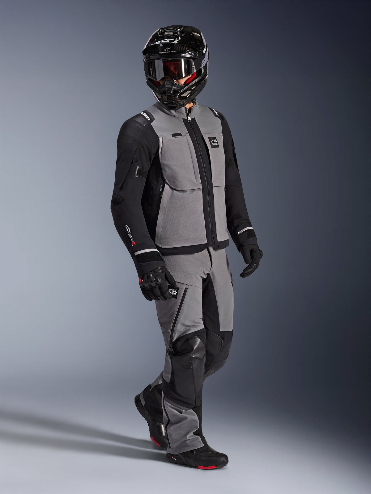 Pilota adventure che indossa la giacca Alpinestars Andes Pro Drystar® XF in grigio scuro e nero, pantaloni adventure coordinati grigi e neri, casco integrale nero con maschera e guanti da moto neri, scatto in studio per attrezzatura professionale da touring a lunga distanza.