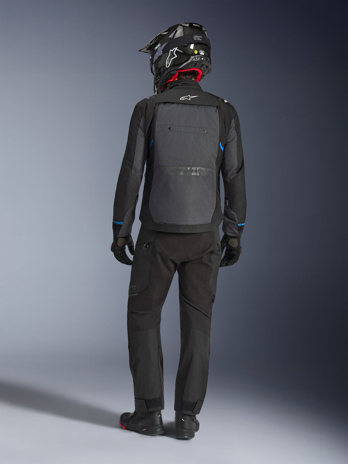 Giacca Alpinestars Cusco Drystar®, nero e grigio scuro, vista posteriore del pilota con abbigliamento adventure touring completo, ampio pannello posteriore grigio con logo Alpinestars, dettagli blu sulle maniche, abbinata a pantaloni neri e casco.