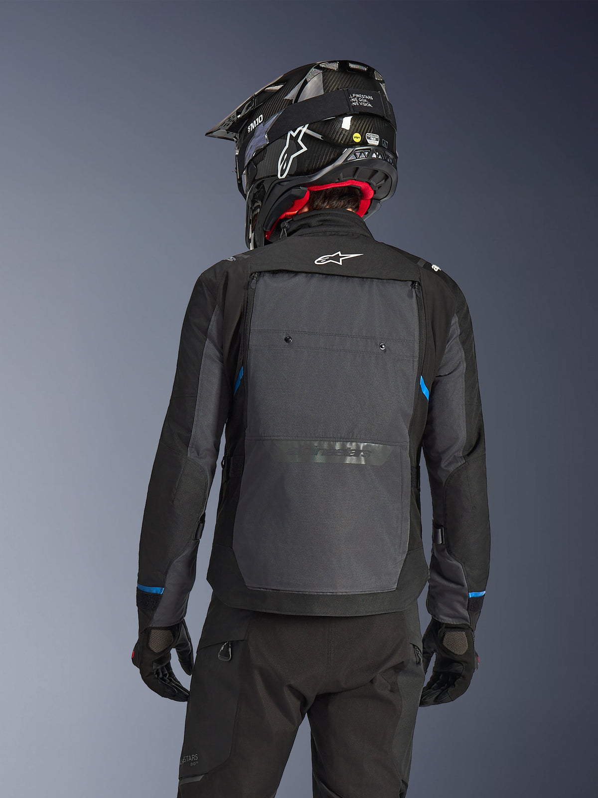 Pilota visto da dietro che indossa la giacca Alpinestars Cusco Drystar® in nero e grigio scuro, con un ampio pannello di ventilazione grigio sulla schiena e logo bianco, abbinata a un casco off-road nero e abbigliamento tecnico nero per l'adventure touring.