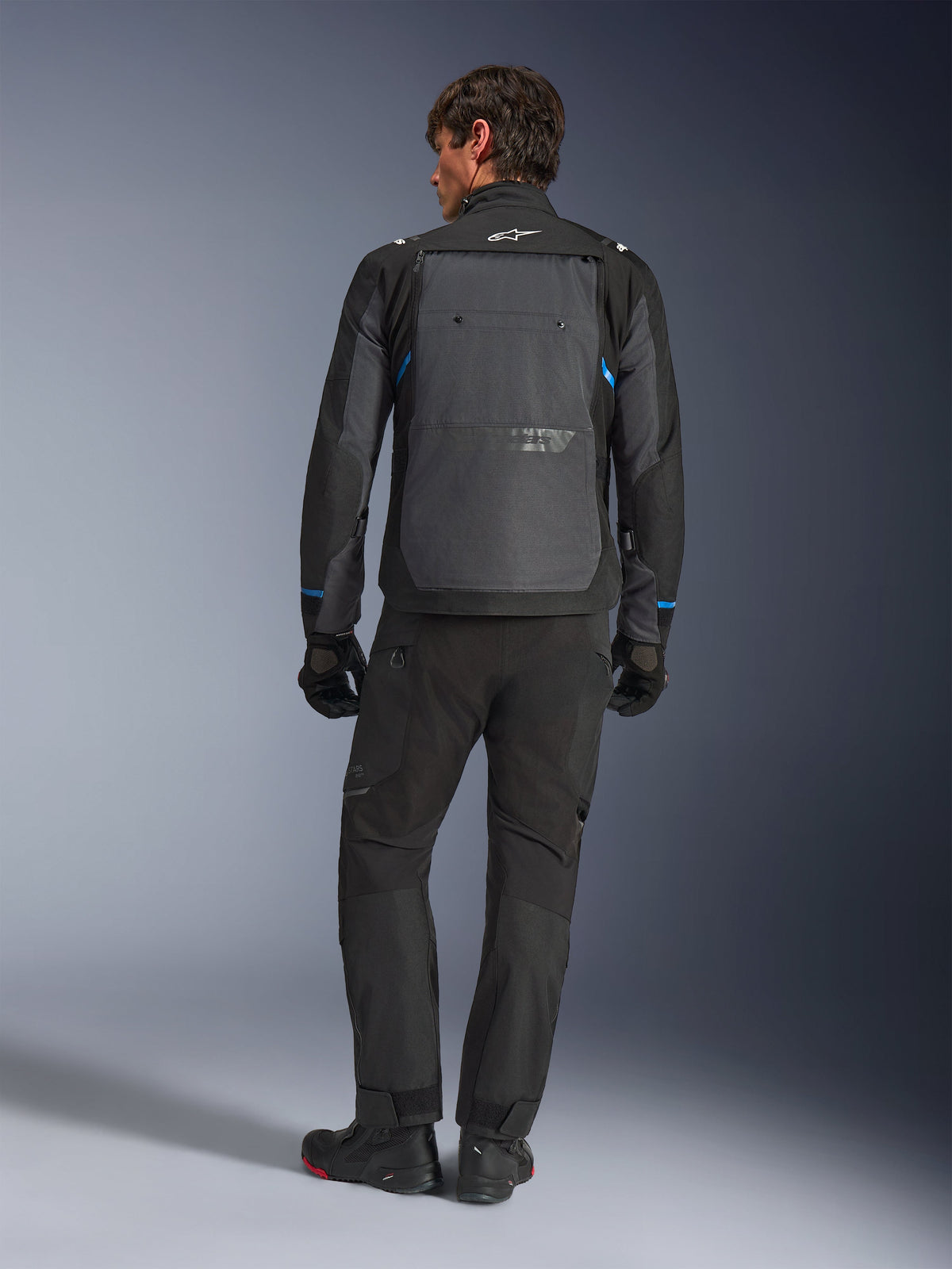 Giacca Alpinestars Cusco Drystar®, nero e grigio scuro con accenti blu, vista posteriore, indossata con pantaloni adventure e guanti neri abbinati, caratterizzata da un ampio pannello posteriore grigio e logo bianco sul colletto, progettata per il touring su strada e l'adventure touring