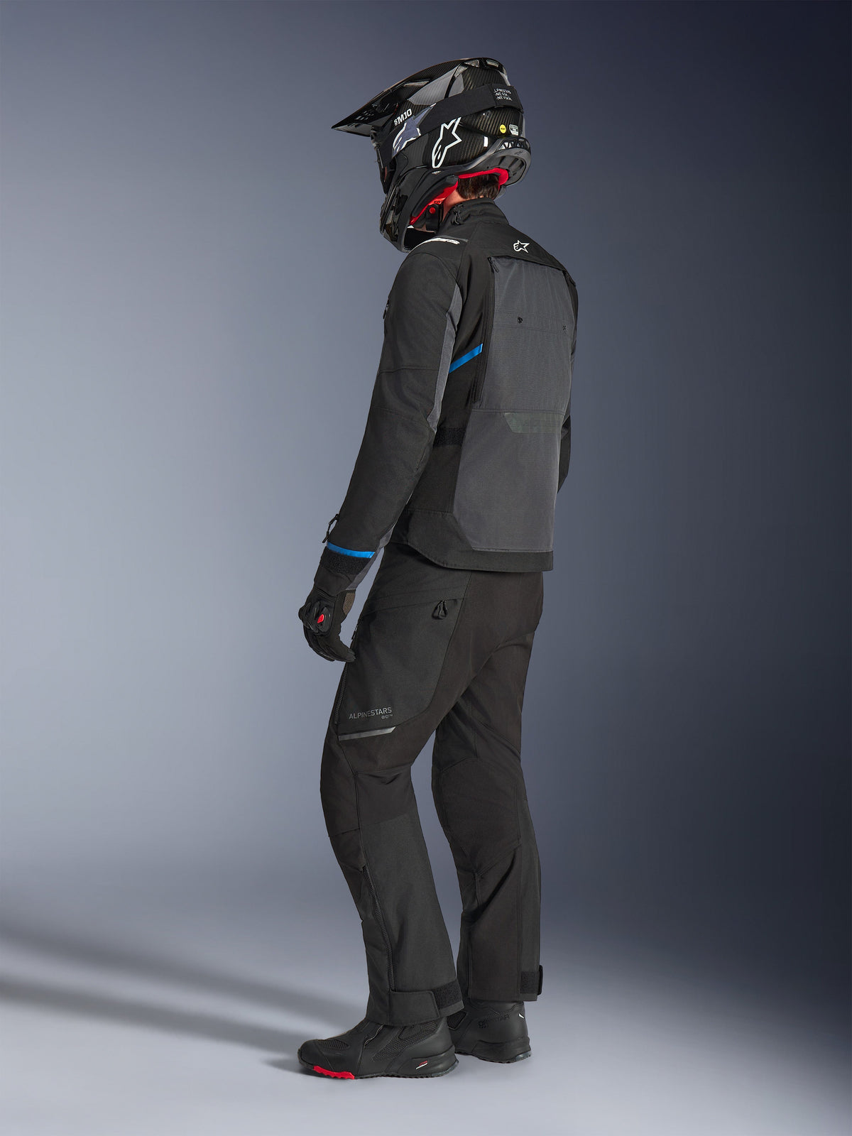 Motociclista che indossa la giacca Alpinestars Cusco Drystar® in nero e grigio scuro con accenti blu, abbinata a pantaloni da moto neri, un casco SM10 in fibra di carbonio e guanti neri, vista posteriore che mostra l'ampio pannello di ventilazione e il design adventure touring.