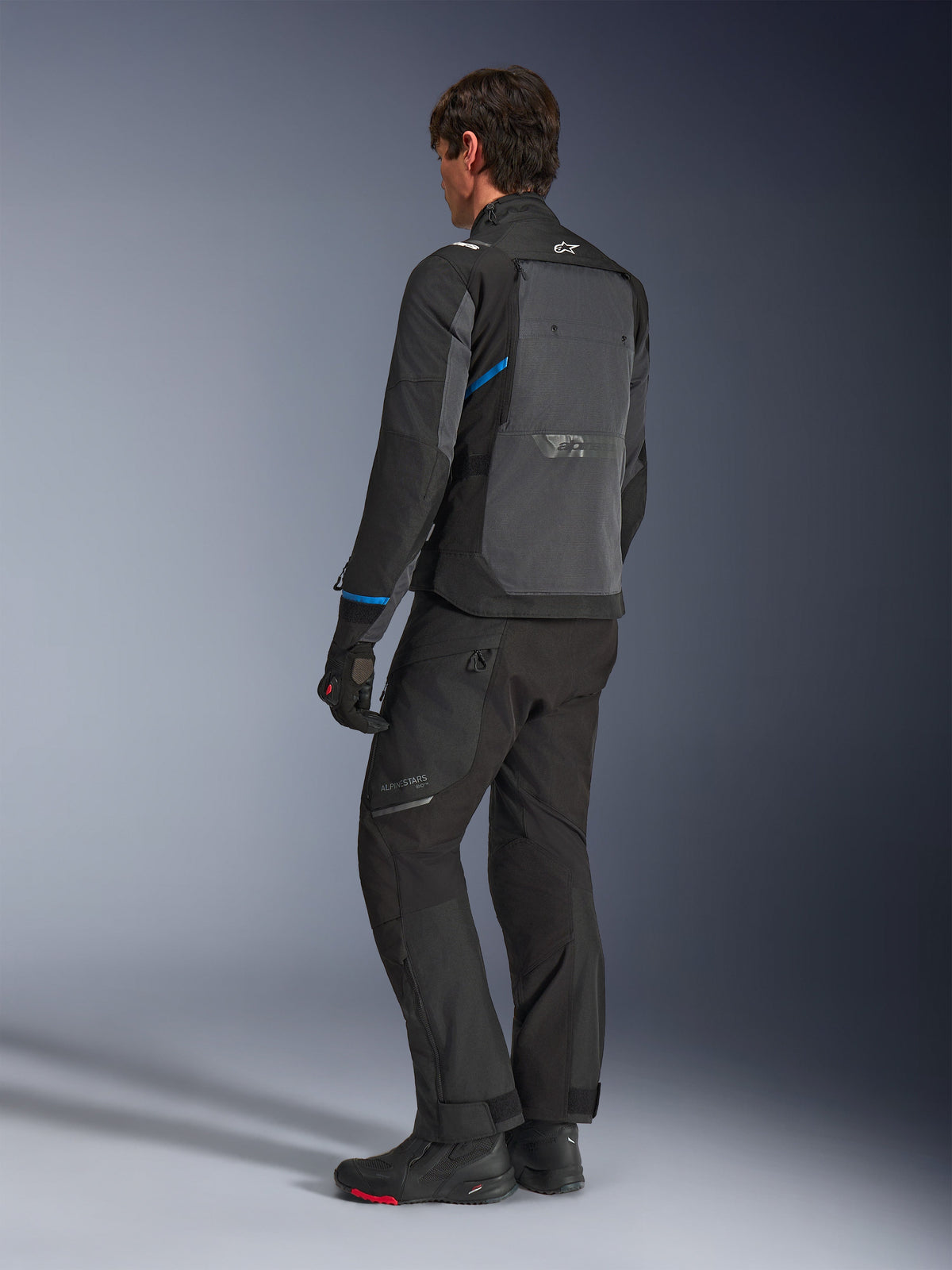 Giacca Alpinestars Cusco Drystar®, nero e grigio scuro, vista posteriore a tre quarti che mostra l'ampio pannello di ventilazione posteriore e dettagli blu sulle maniche, indossata con pantaloni e stivali da moto neri per un outfit completo da touring.