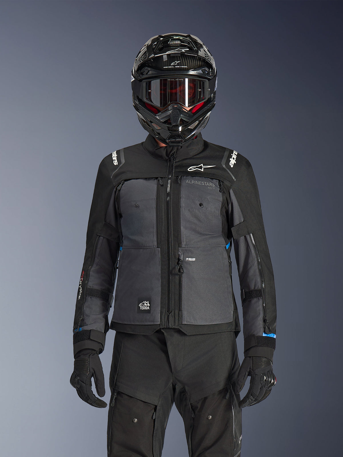 Giacca Alpinestars Cusco Drystar®, Nero e Grigio Scuro, giacca da moto adventure touring indossata con pantaloni tecnici neri abbinati e un casco dual-sport, caratterizzata da un design bicolore con pannelli in ripstop e branding Alpinestars