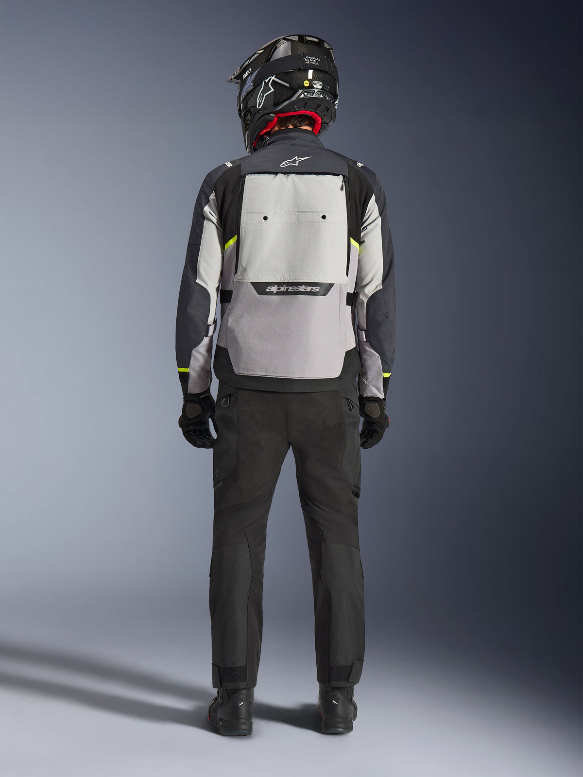 Motociclista visto da dietro che indossa la giacca Alpinestars Cusco Drystar® in Ice Gray, Mid Gray e Deep Gray, con un ampio pannello di ventilazione posteriore e logo Alpinestars, abbinata a pantaloni da moto neri e un casco nero, abbigliamento da moto adventure touring
