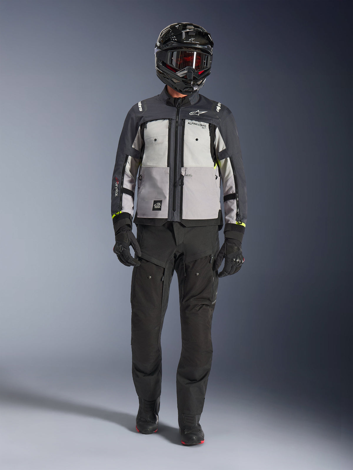 Giacca Alpinestars Cusco Drystar®, Ice Gray Mid Gray Deep Gray, giacca da moto adventure indossata con pantaloni da guida neri, guanti neri e casco nero con occhiali, caratterizzata da pannelli grigi color-block e dettagli tecnici per il touring su strada e adventure.