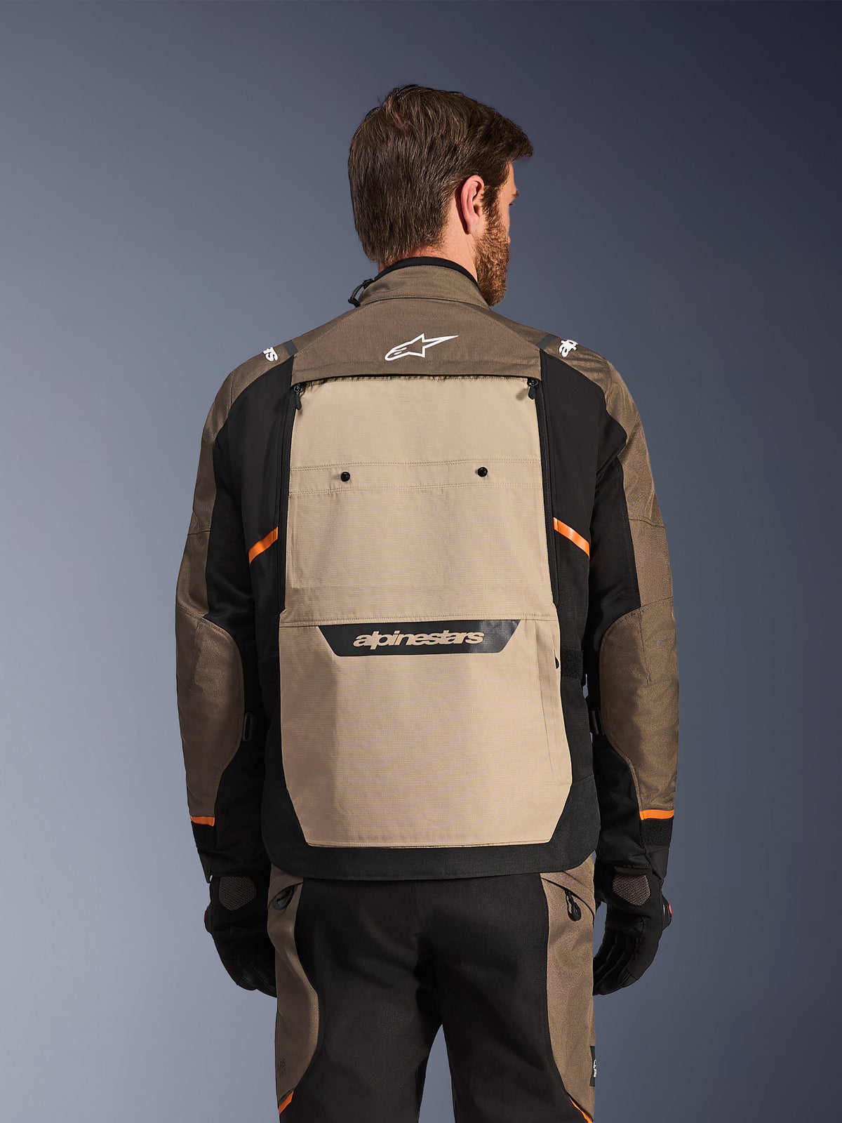 Giacca Alpinestars Cusco Drystar®, Walnut Aluminum Black, vista posteriore, ampio pannello posteriore color sabbia con logo nero, accenti arancioni, indossata con pantaloni e guanti coordinati, progettata per l'adventure touring.