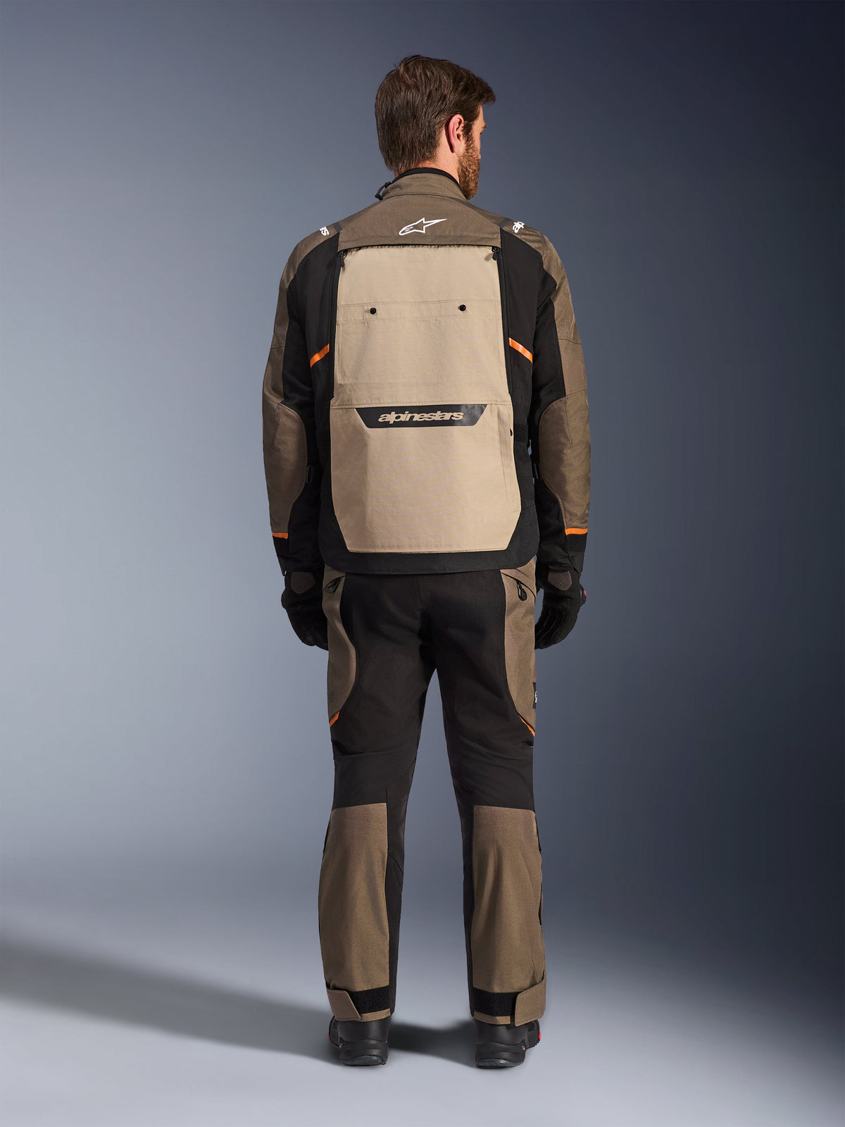 Alpinestars Giacca Cusco Drystar®, vista posteriore, Walnut Aluminum Black, abbigliamento tecnico da adventure touring, ampio pannello di ventilazione posteriore color sabbia con logo nero, accenti arancioni, indossata con pantaloni e guanti coordinati.