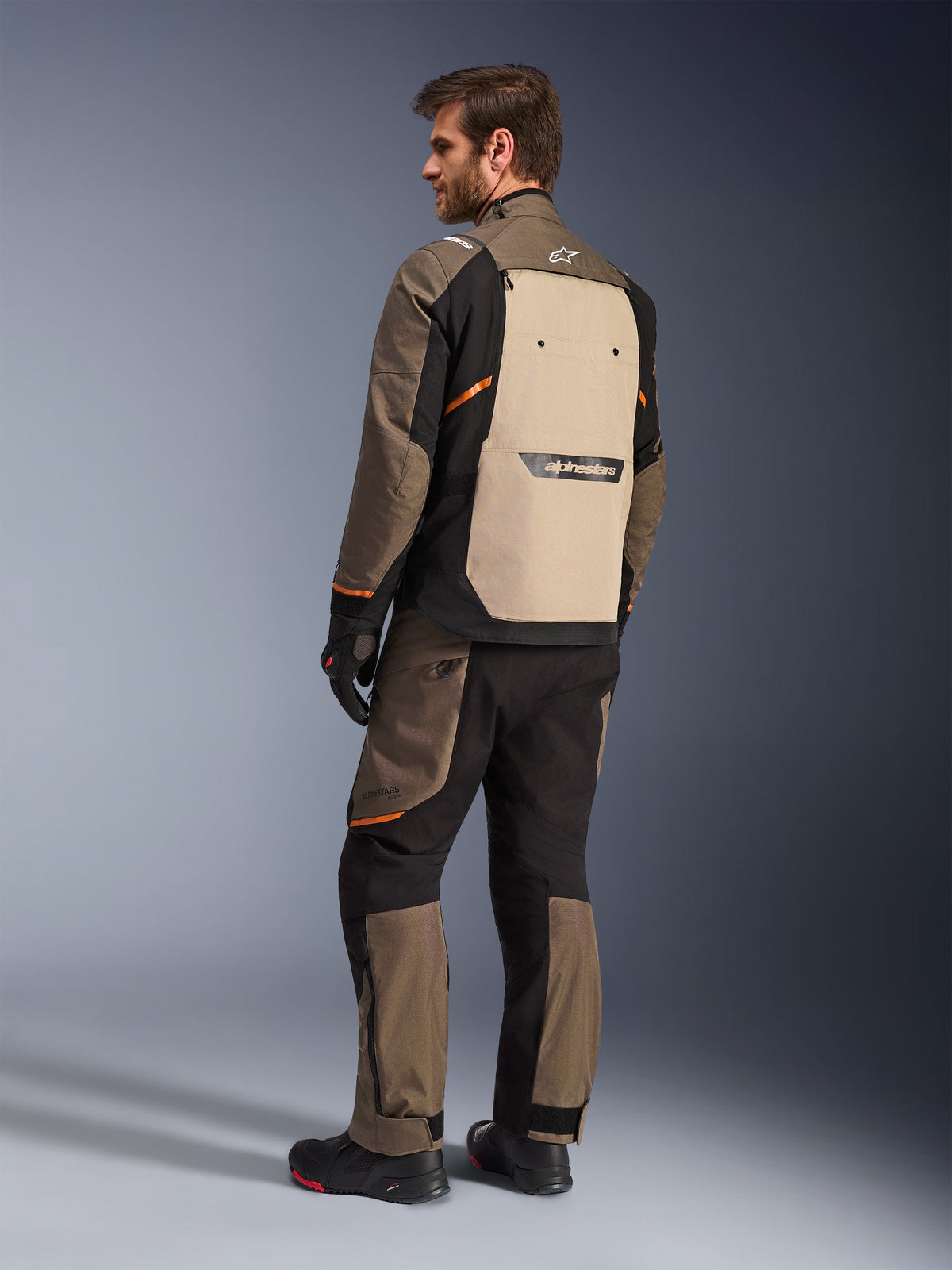 Giacca Alpinestars Cusco Drystar® in Walnut Aluminum Black, vista posteriore che mostra il pannello posteriore beige e dettagli arancioni, indossata con pantaloni da moto coordinati, guanti e stivali neri, abbigliamento professionale da adventure touring