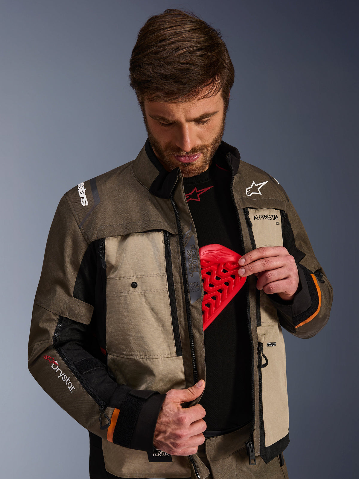Giacca Alpinestars Cusco Drystar®, Walnut Aluminum Black, indossata da un uomo che mostra la tasca per la protezione del torace con un inserto protettivo rosso, giacca da moto in tessuto con prese d'aria con cerniera e loghi per adventure touring.