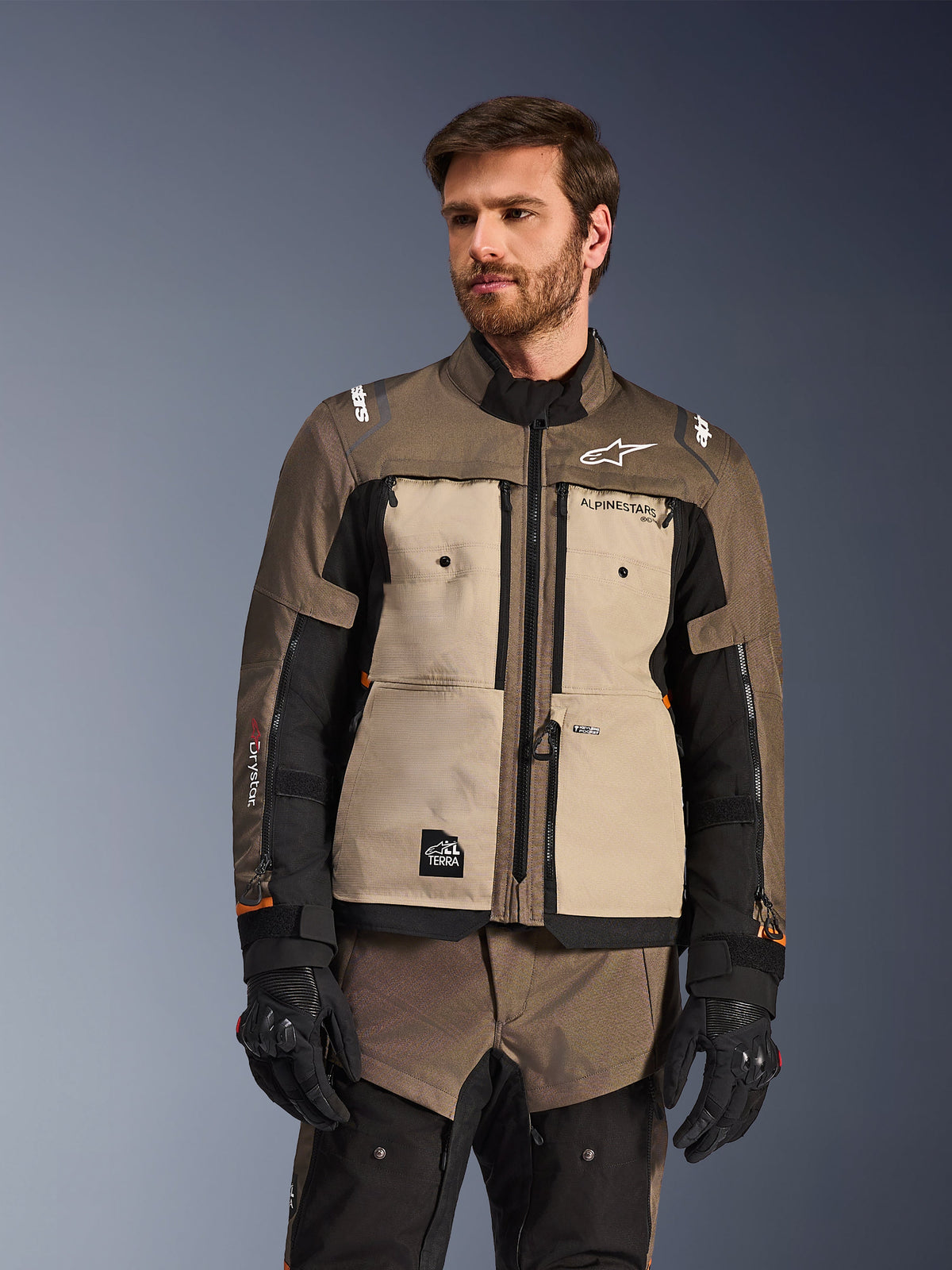 Giacca Alpinestars Cusco Drystar®, giacca da moto adventure, Walnut Aluminum Black, indossata da un modello con guanti neri, con pannelli sul petto color sabbia e maniche nere con marchio Alpinestars e toppa All Terra, abbigliamento protettivo da touring per ogni condizione atmosferica