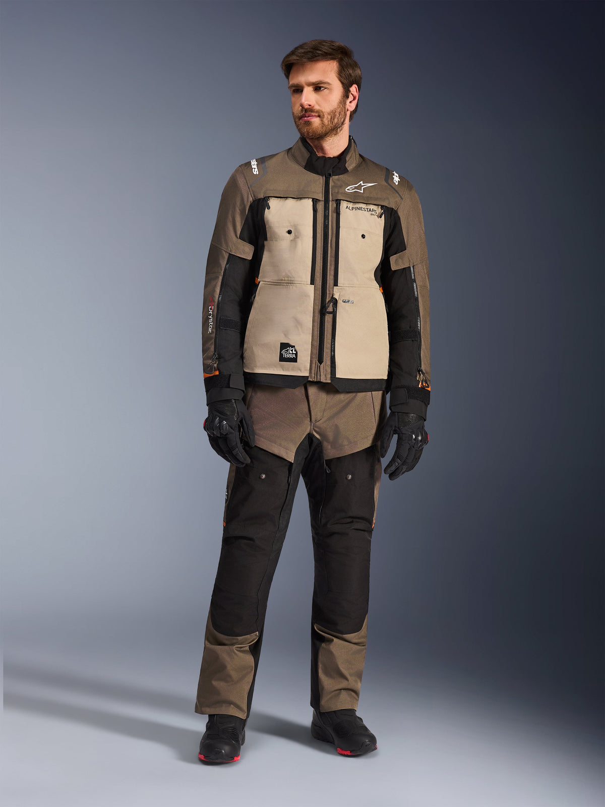 Giacca Alpinestars Cusco Drystar® in Walnut Aluminum Black, indossata da un motociclista con pantaloni adventure touring abbinati, guanti e stivali neri, con pannelli sul petto color sabbia, maniche nere con sottili accenti arancioni e cerniere di ventilazione funzionali per la guida su strada.