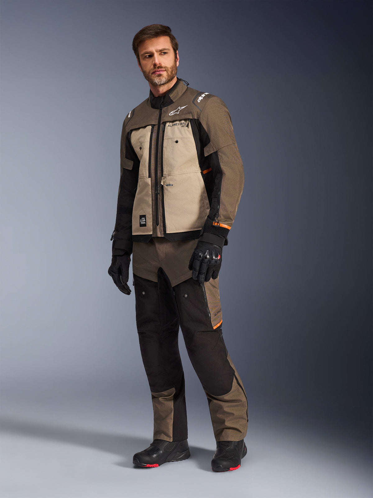 Giacca Alpinestars Cusco Drystar® in Walnut Aluminum Black, indossata da un modello con pantaloni e guanti da moto coordinati, design adventure-touring con pannelli sul petto color sabbia e accenti neri.