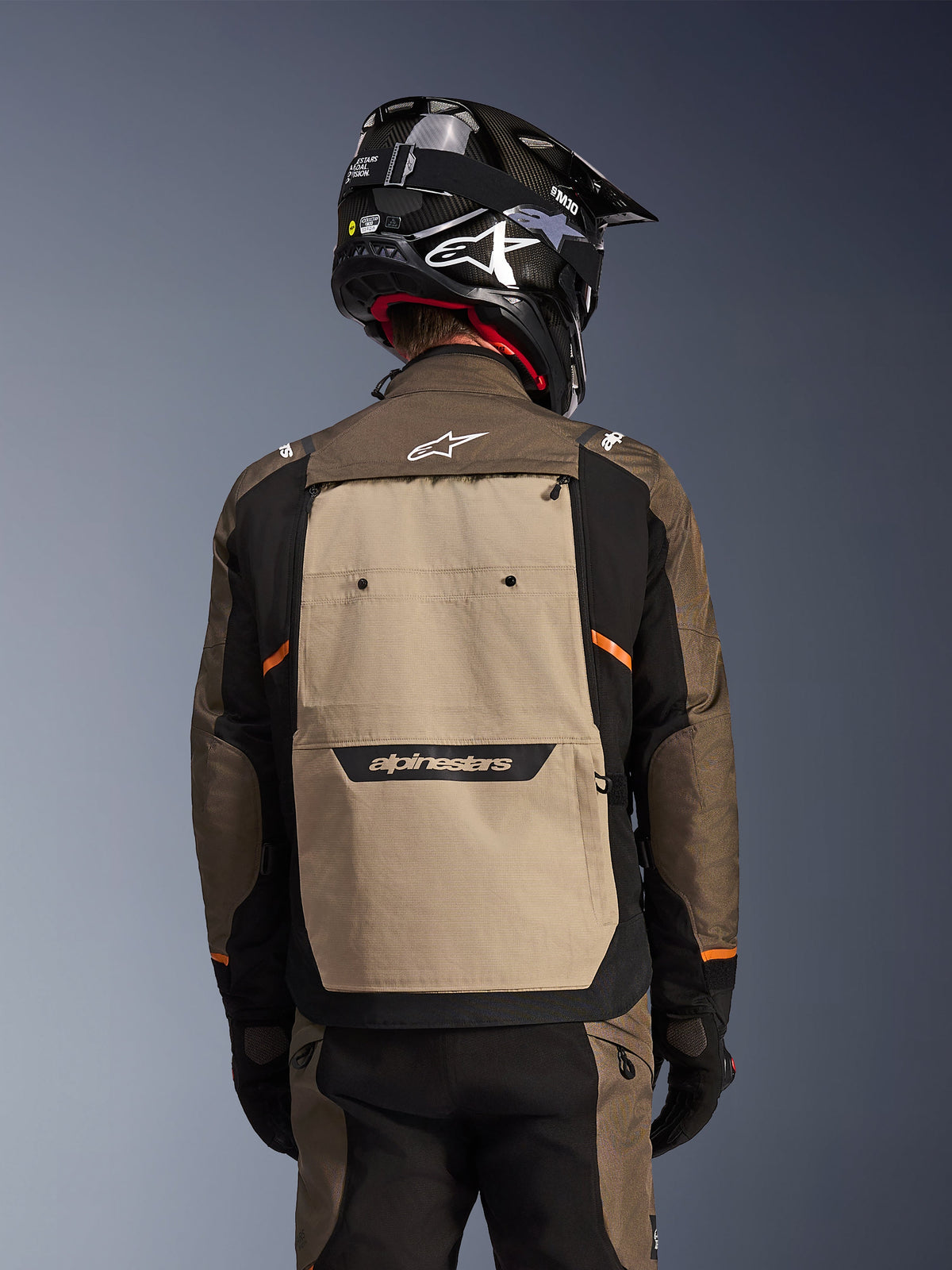 Vista posteriore della giacca Alpinestars Cusco Drystar® in Walnut Aluminum Black, giacca da moto adventure touring con ampio pannello posteriore color sabbia e logo Alpinestars, maniche nere con accenti arancioni, mostrata con un casco nero stile carbonio